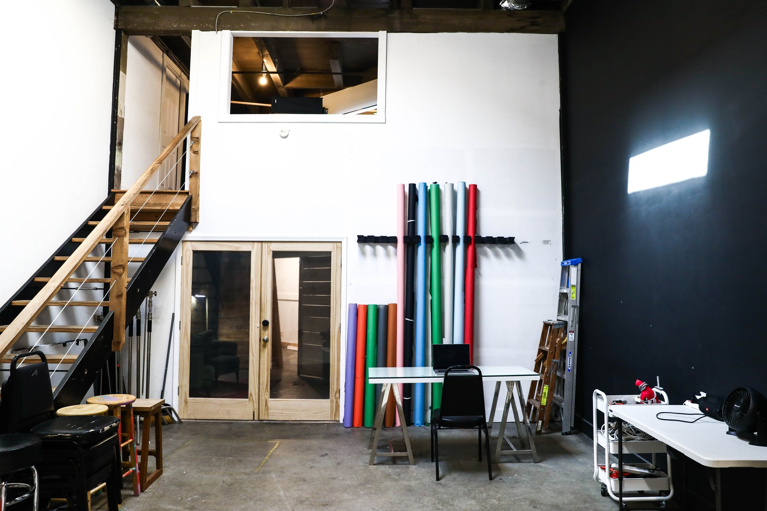 Interior of an industrial-style room with a wooden staircase to a loft area, colorful foam tubes against a white wall, a small wooden ladder, black chairs stacked, a white table with a laptop, and a white cart with equipment, all illuminated by overh