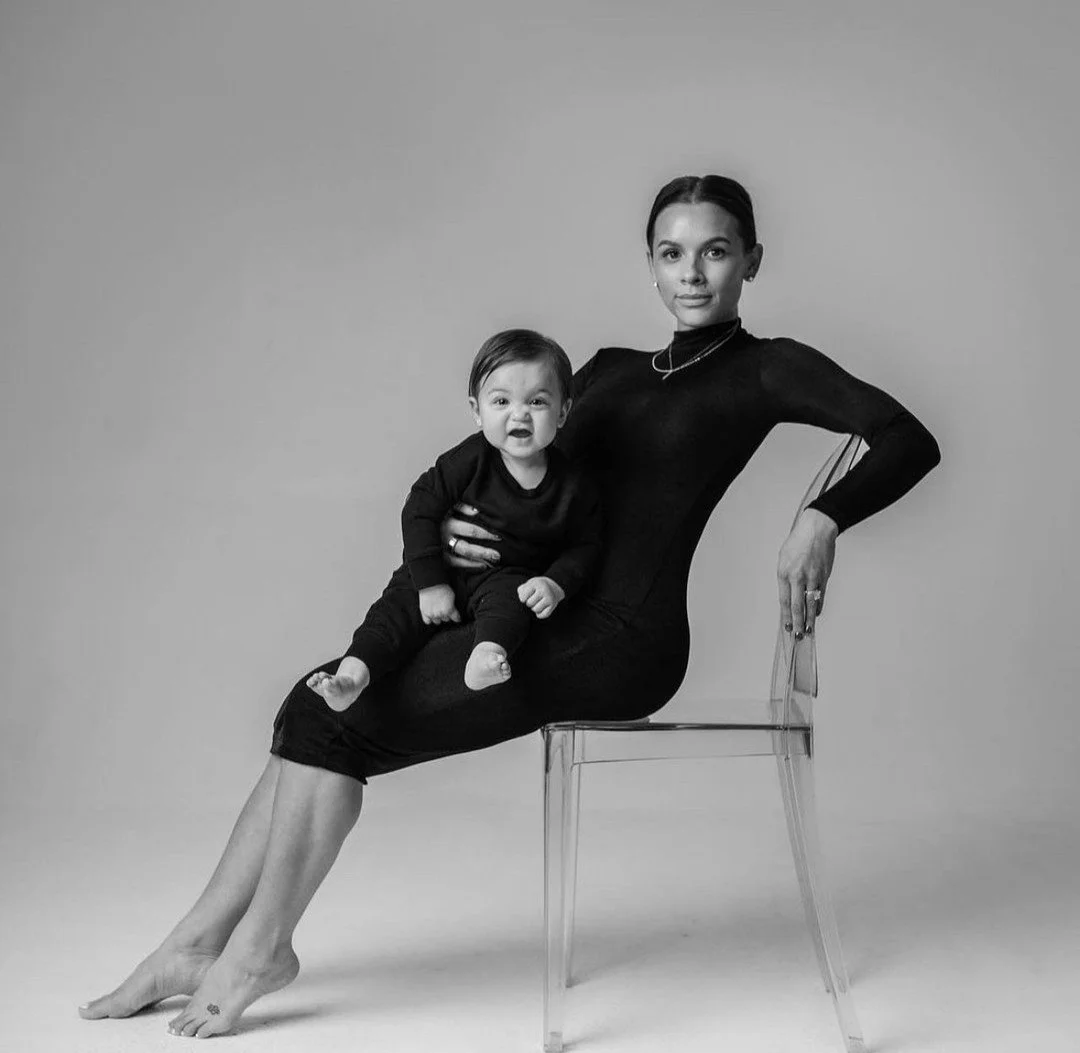 A woman sitting on a transparent chair holding a young child on her lap. The woman is wearing a form-fitting dress, and the child is dressed in dark clothing. Both are looking at the camera. The image is black and white.