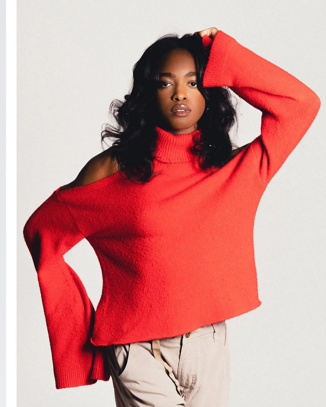 A woman wearing a red off-the-shoulder sweater with a turtleneck, posing with one hand on her head against a plain white background.