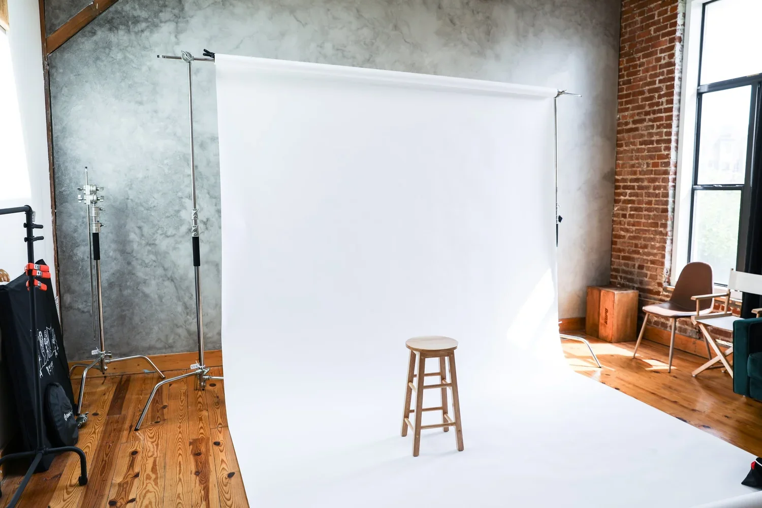 Indoor photography studio with a white backdrop, a wooden stool in center, and multiple chairs nearby on wooden floor, brick and concrete wall with large windows.