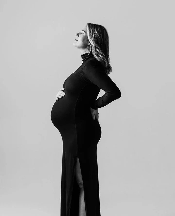 Black and white photo of a pregnant woman in a long dress with a slit, standing sideways with eyes closed and hands on her belly and lower back, against a plain background.
