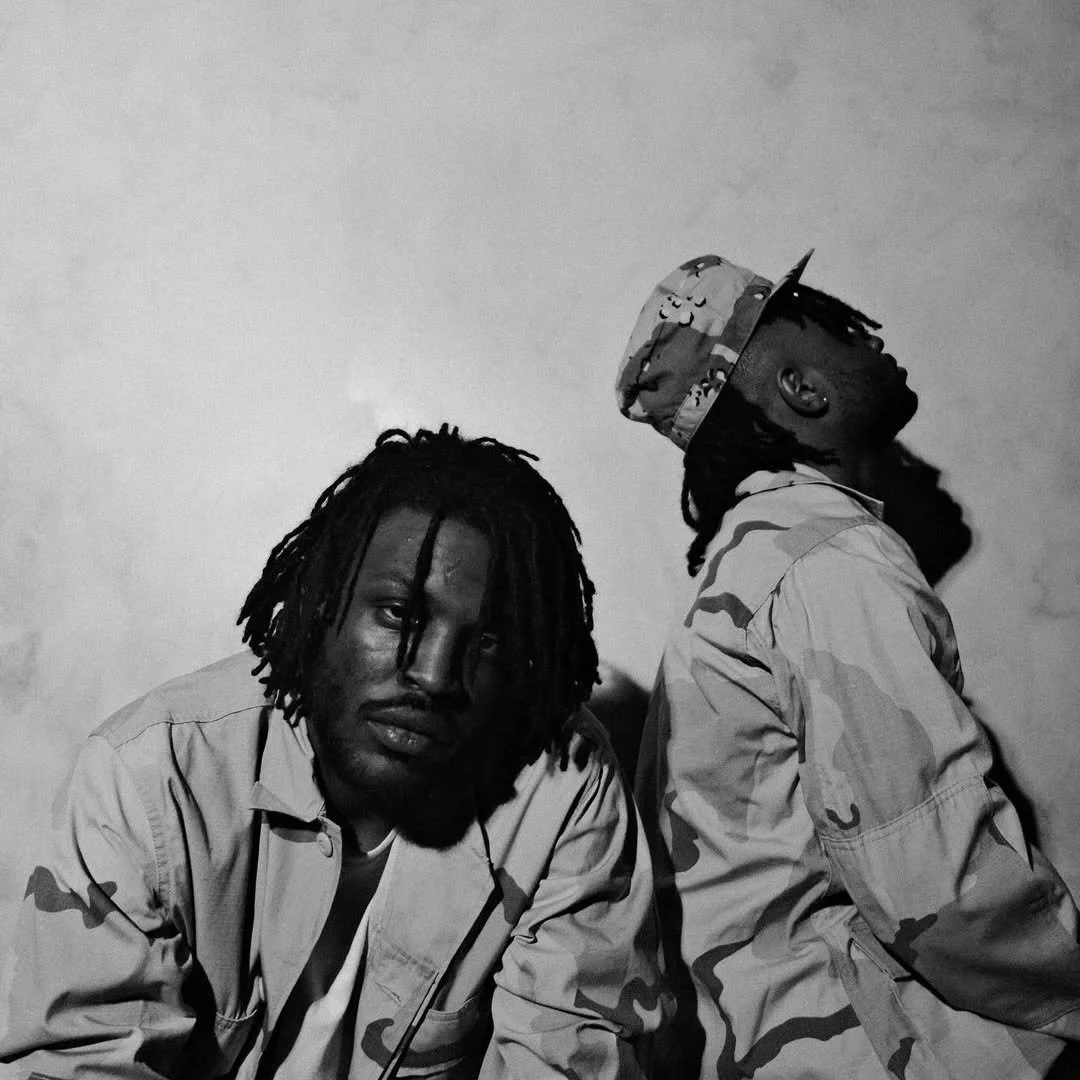 Two men with dreadlocks wearing military-style camouflage jackets and one wearing a camouflage cap, against a plain wall.