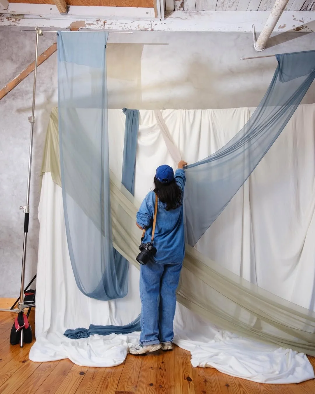 A person in blue clothing and a blue baseball cap is arranging sheer blue and light beige fabric panels on a white backdrop in an art installation or photography studio, with some fabric draped on the wooden floor.