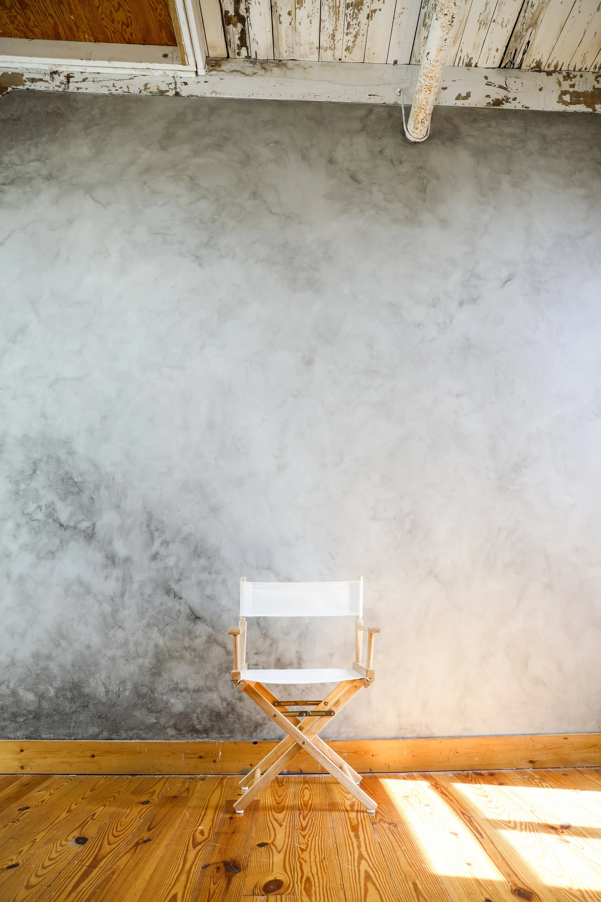 A minimalist indoor scene with a white folding chair with wooden armrests and legs, placed against a textured gray wall and natural wooden floor, with sunlight casting shadows on the floor.
