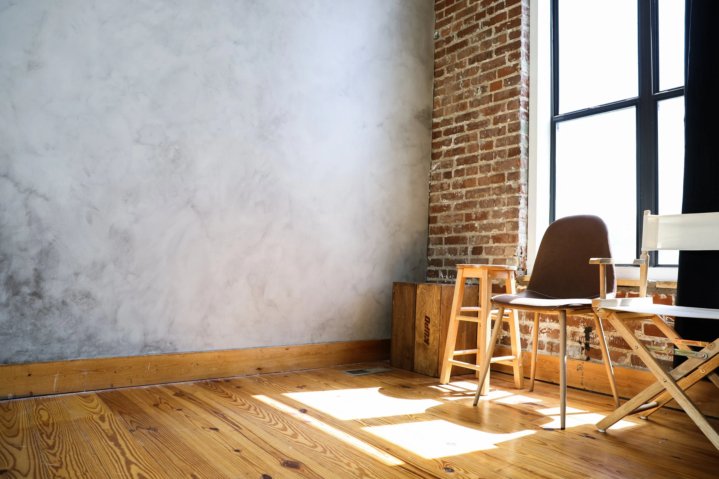 Sunlight coming through a large window into a room with a wooden floor, a brown chair, a small wooden stool, and a wooden box against an exposed brick wall.