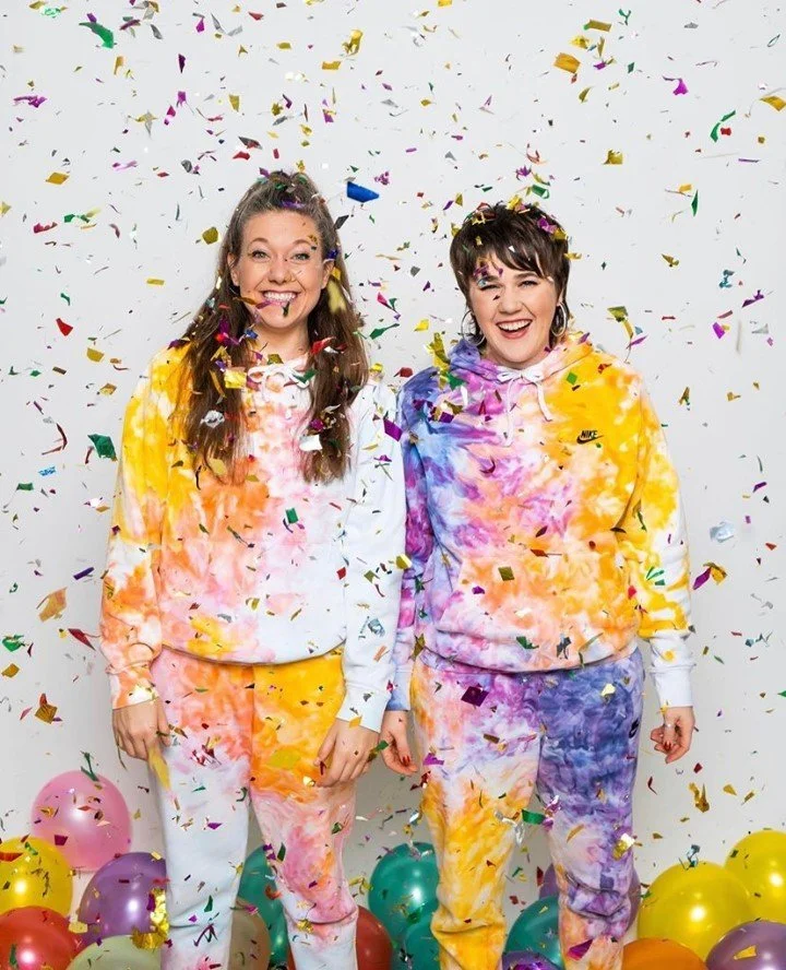 Two women celebrating with colorful confetti in the air, wearing tie-dye hoodies and sweatpants, surrounded by balloons at a festive event.