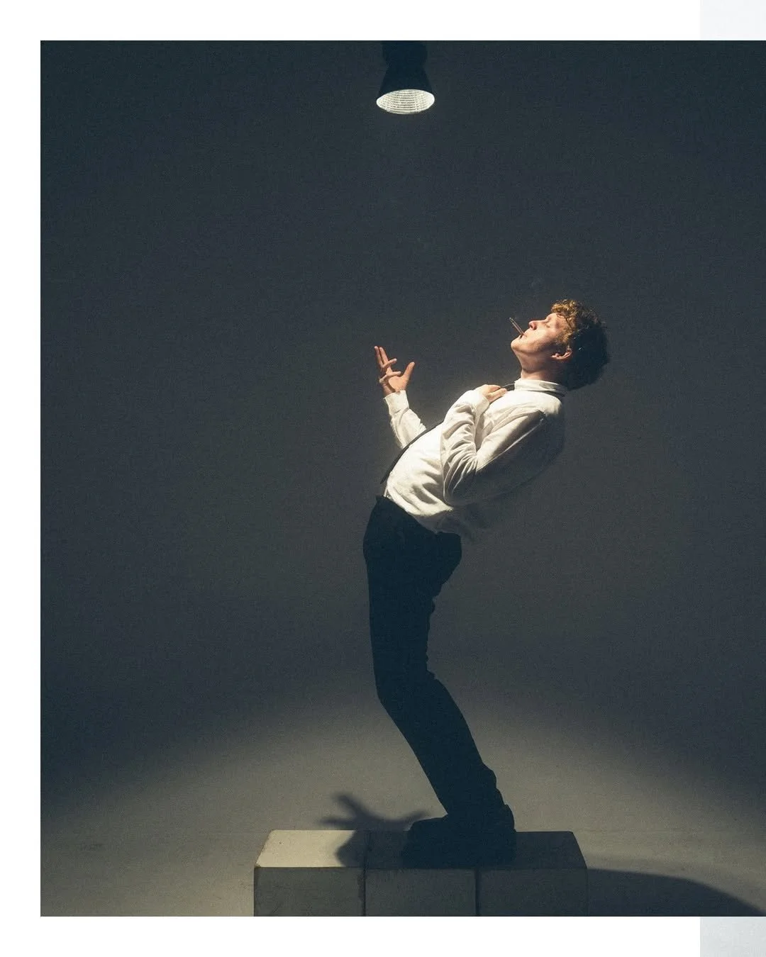 A man in formal attire standing on a block, posing with his head tilted back, smoking a cigarette, and looking up at a hanging light fixture in a dark room.