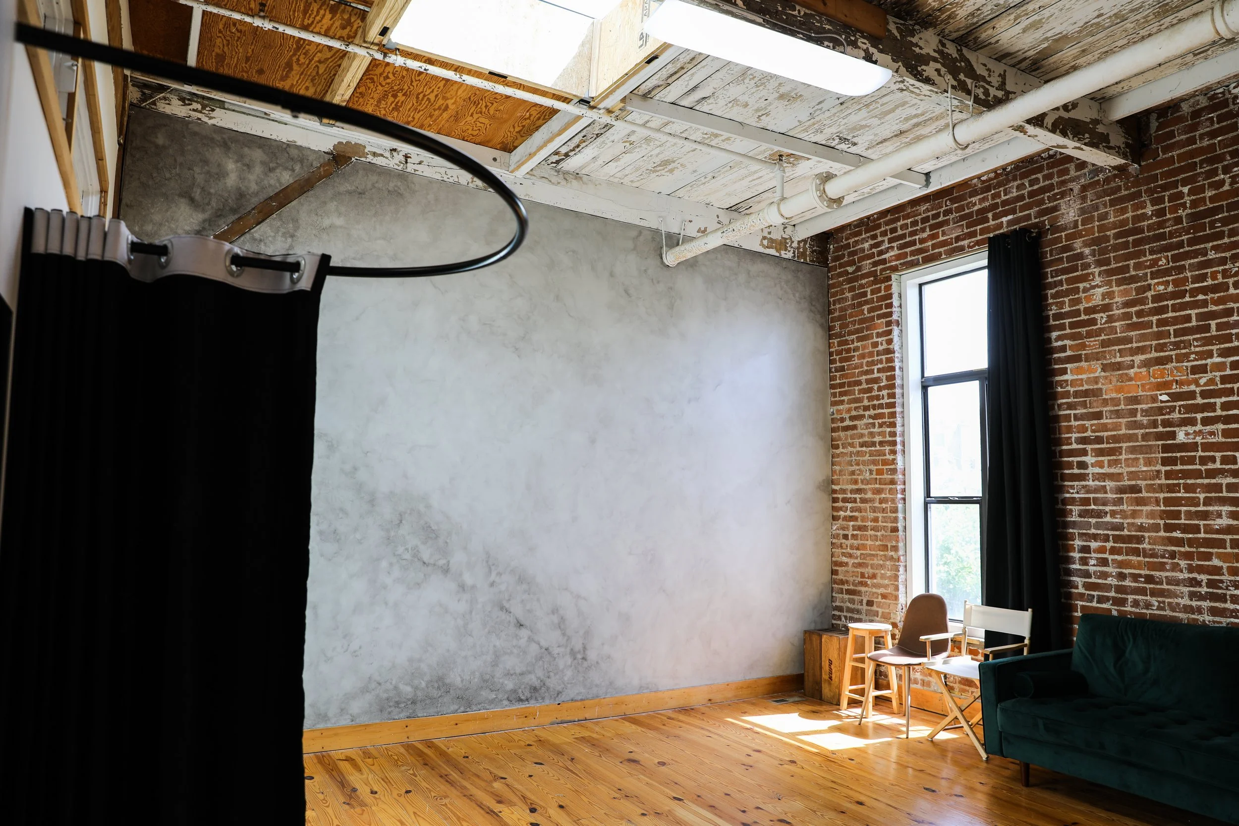 Interior of an industrial-style room with exposed brick and concrete walls, large window with black curtains, wooden floor, and a few pieces of furniture including chairs and a green velvet sofa.