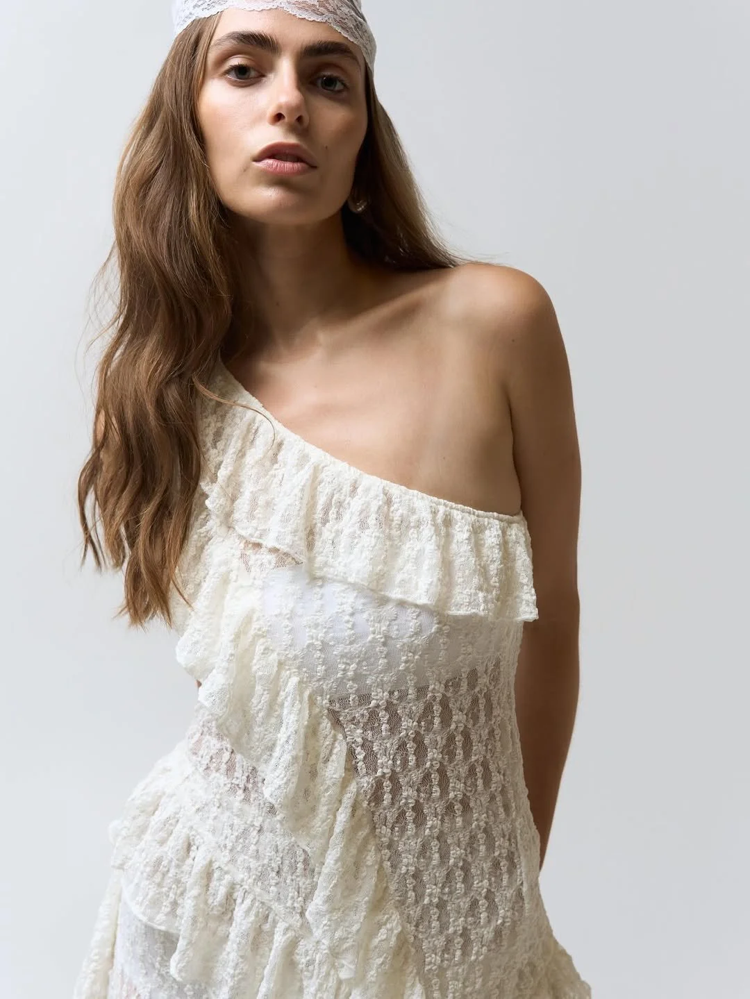 A woman with long brown hair wearing a lace headband and a one-shoulder cream-colored lace dress.