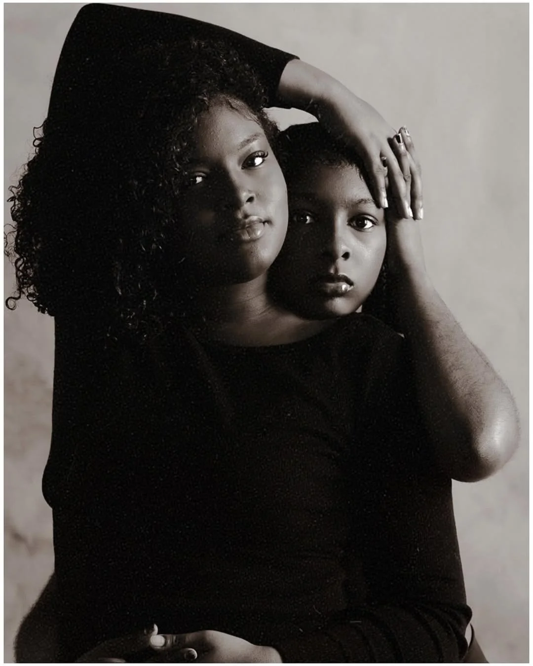 Black and white portrait of two women with curly hair, one resting her arm on the other's head, both looking directly at the camera with serious expressions.