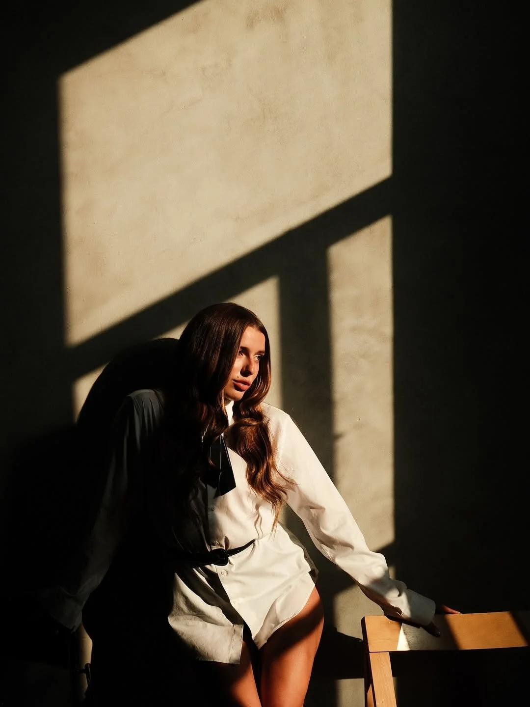 A woman with long, wavy brown hair sitting on a chair with sunlight casting shadows through a window onto the wall behind her.