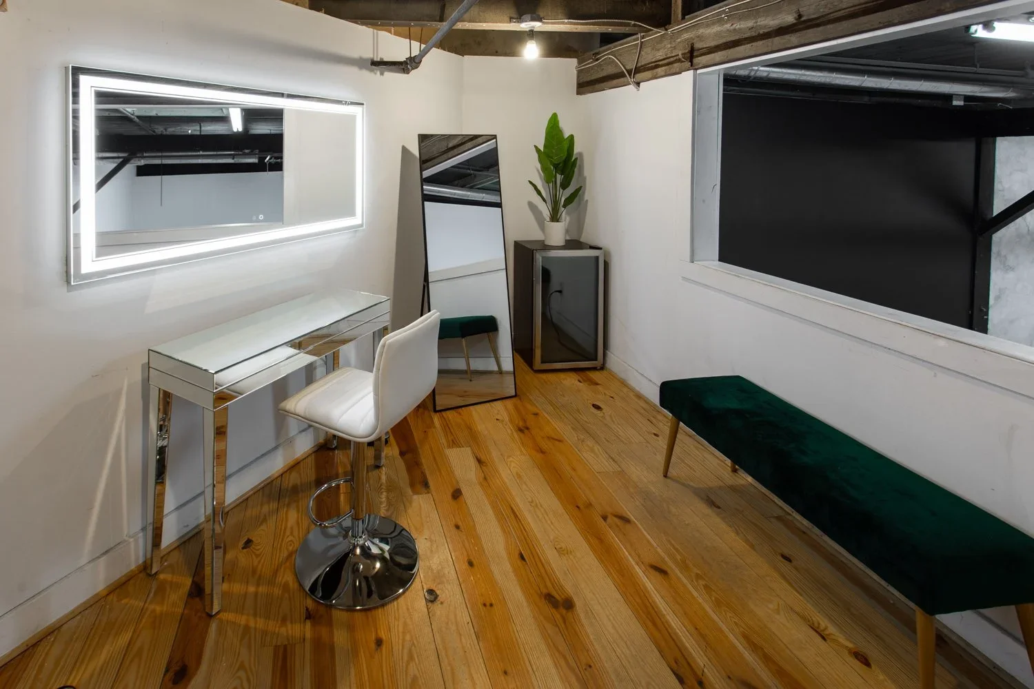 Interior view of a modern room with wooden floor, white walls, and ceiling. Contains a white chair at a mirrored vanity table, a large illuminated mirror on the wall, a full-length mirror, a small cabinet with a potted plant, a bench with dark green 