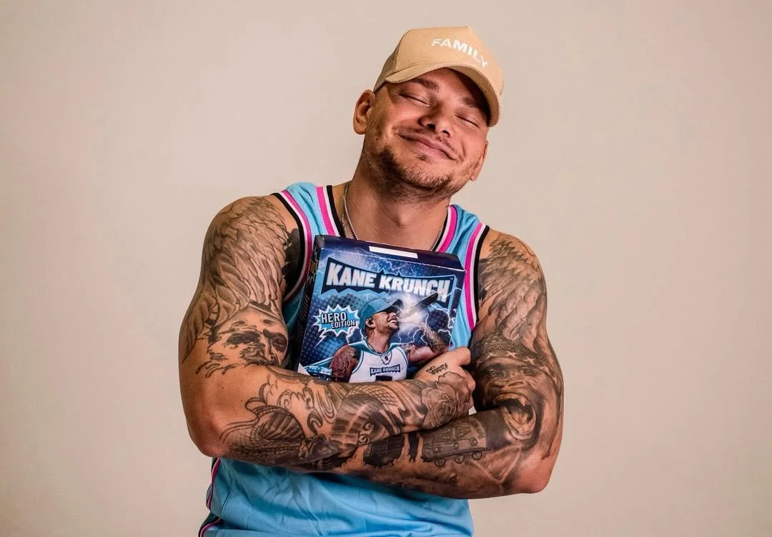 A smiling man wearing a beige family cap and a colorful basketball jersey, holding a snack box labeled 'Kane Krunch Hero Edition' while standing against a plain background.