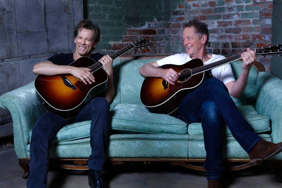 Two men sitting on a vintage light blue couch, playing acoustic guitars and smiling at each other, in an industrial-style room with exposed brick and concrete walls.