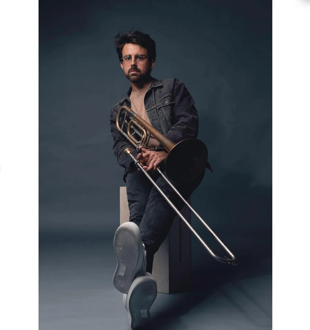 A man with glasses and dark hair, wearing a denim jacket and gray shirt, sitting on a white block, holding a trombone, against a dark gray background.