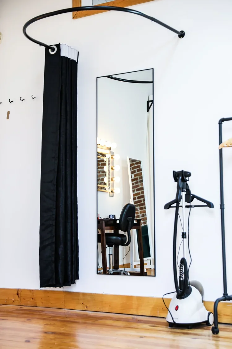 Full-length mirror reflecting a makeup station with a mirror surrounded by lights, a makeup chair, brick wall, and a black curtain, with a garment steamer on the floor near the wall.