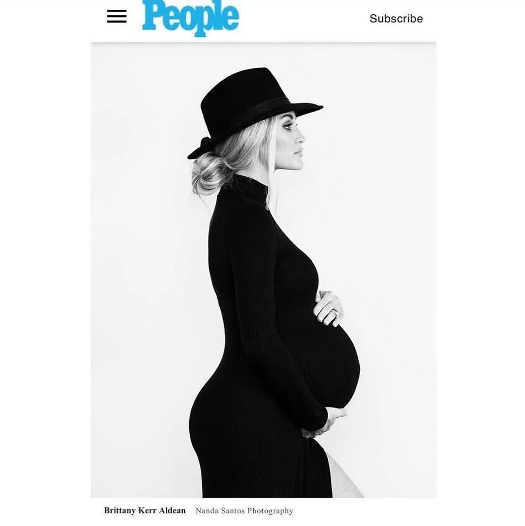 Black and white photograph of a pregnant woman in profile, wearing a fitted black dress and a wide-brimmed black hat, standing against a plain white background.