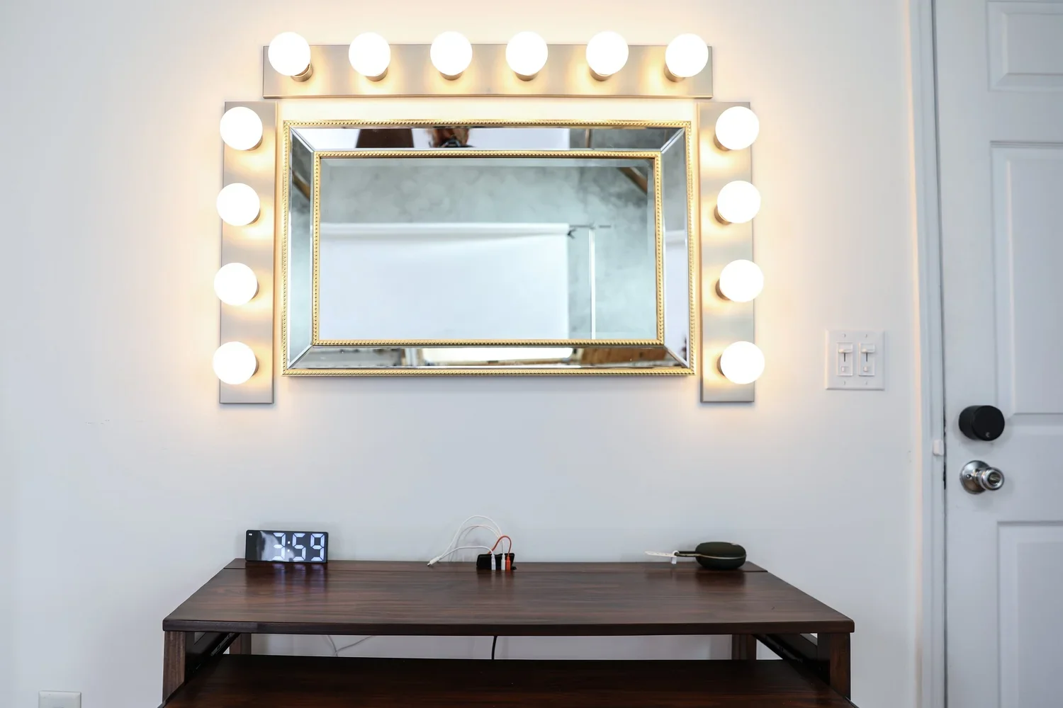 A bedroom vanity mirror with lights, a wooden table underneath, a digital clock displaying 3:59, and some electronic cords and devices on the table. The mirror has a gold trim and is surrounded by light bulbs. There is a door with a black knob on the