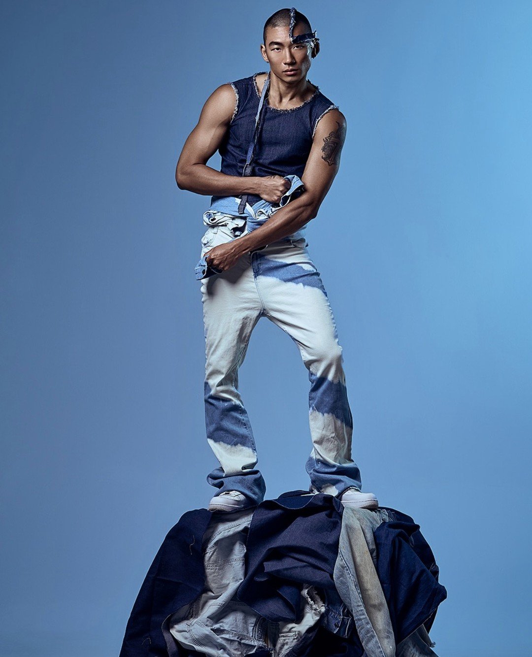 A young man standing on top of a pile of clothes against a blue background, dressed in a ripped sleeveless denim shirt and light-washed jeans with patches, with a blue bandana tied on his head.