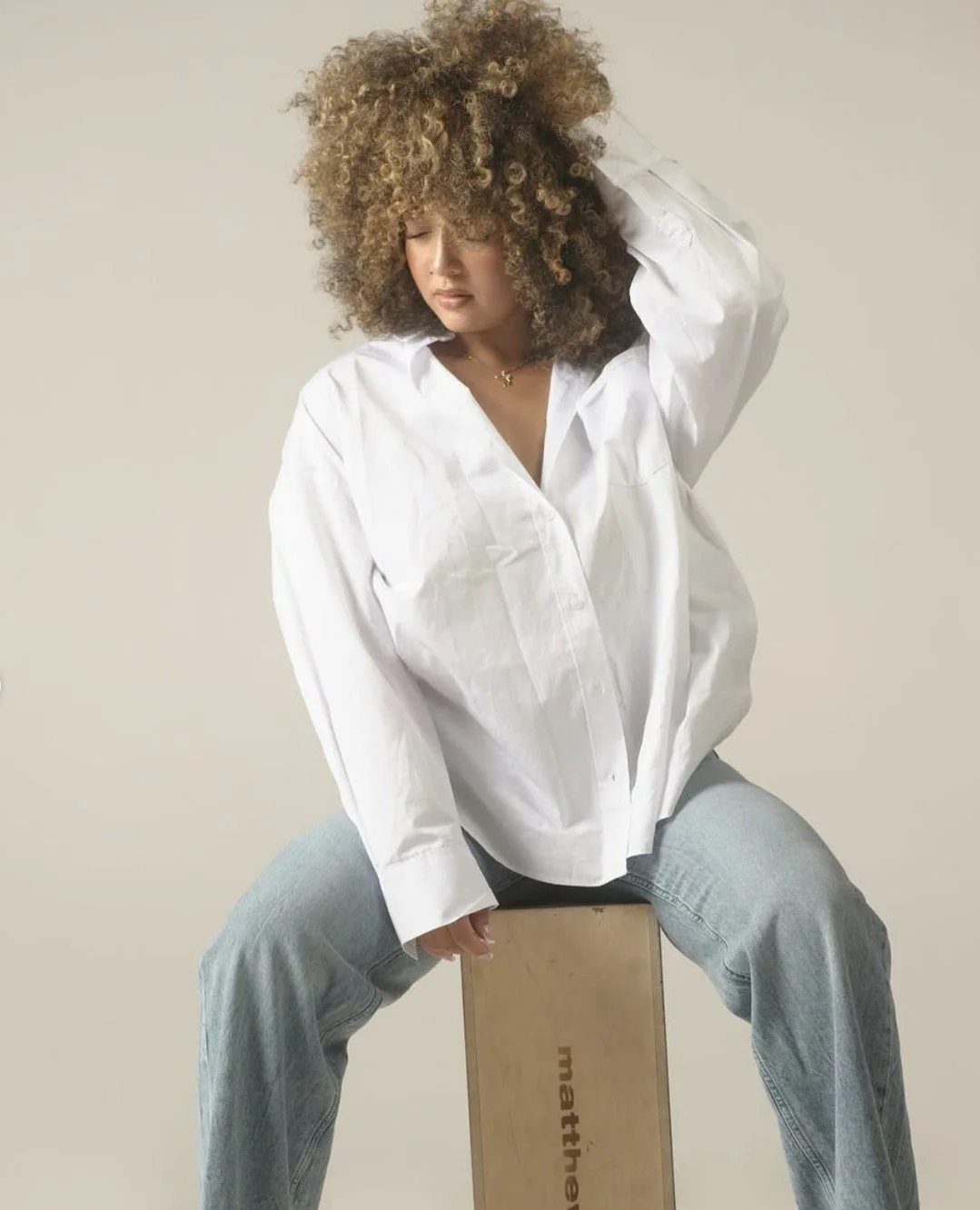 A woman with curly hair wearing an oversized white shirt and light blue jeans, sitting on a wooden box with her eyes closed and one hand on her head.