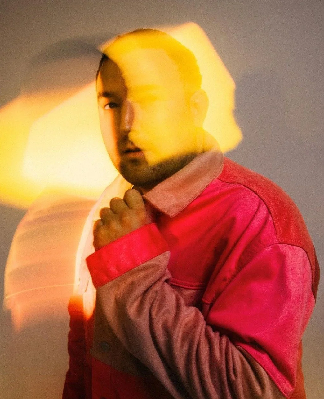 A man wearing a pink and beige jacket with red accents, posing with his hand near his chin, facing the camera. The photograph has a motion blur effect with yellow light streaks in the background.