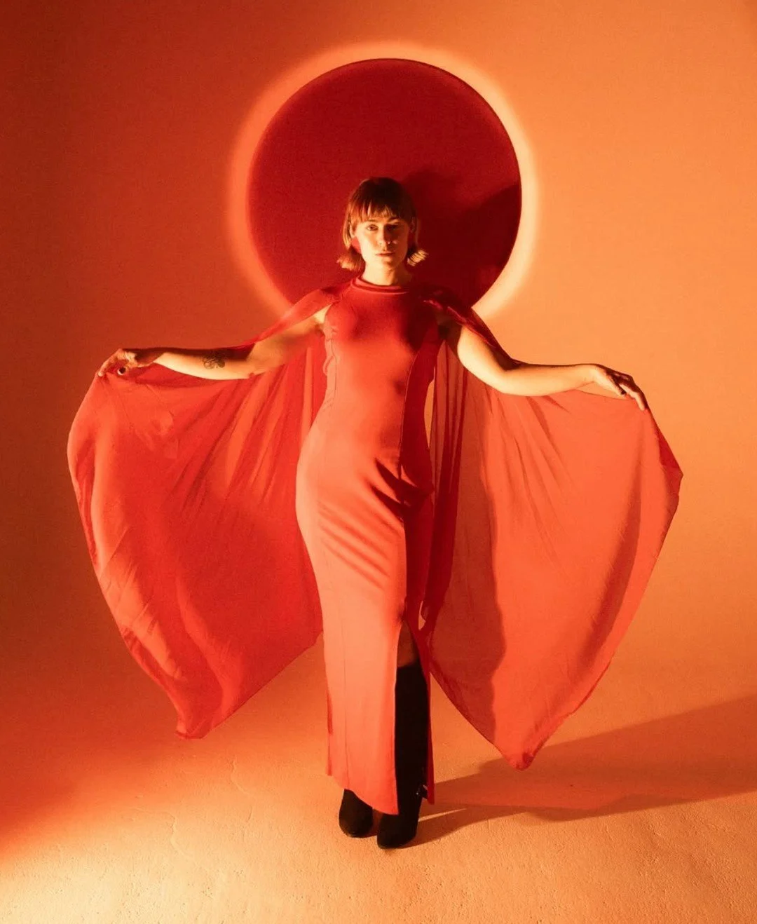 A woman in a long red form-fitting dress with flowing fabric extending from her arms, standing against a reddish-orange background with a glowing circular light behind her head.