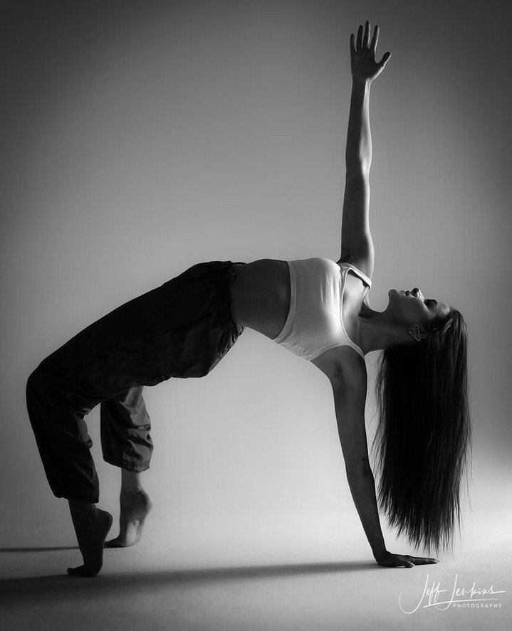 A woman performing a yoga pose balancing on her hands and feet, with one arm reaching upward, in a studio with a plain background.