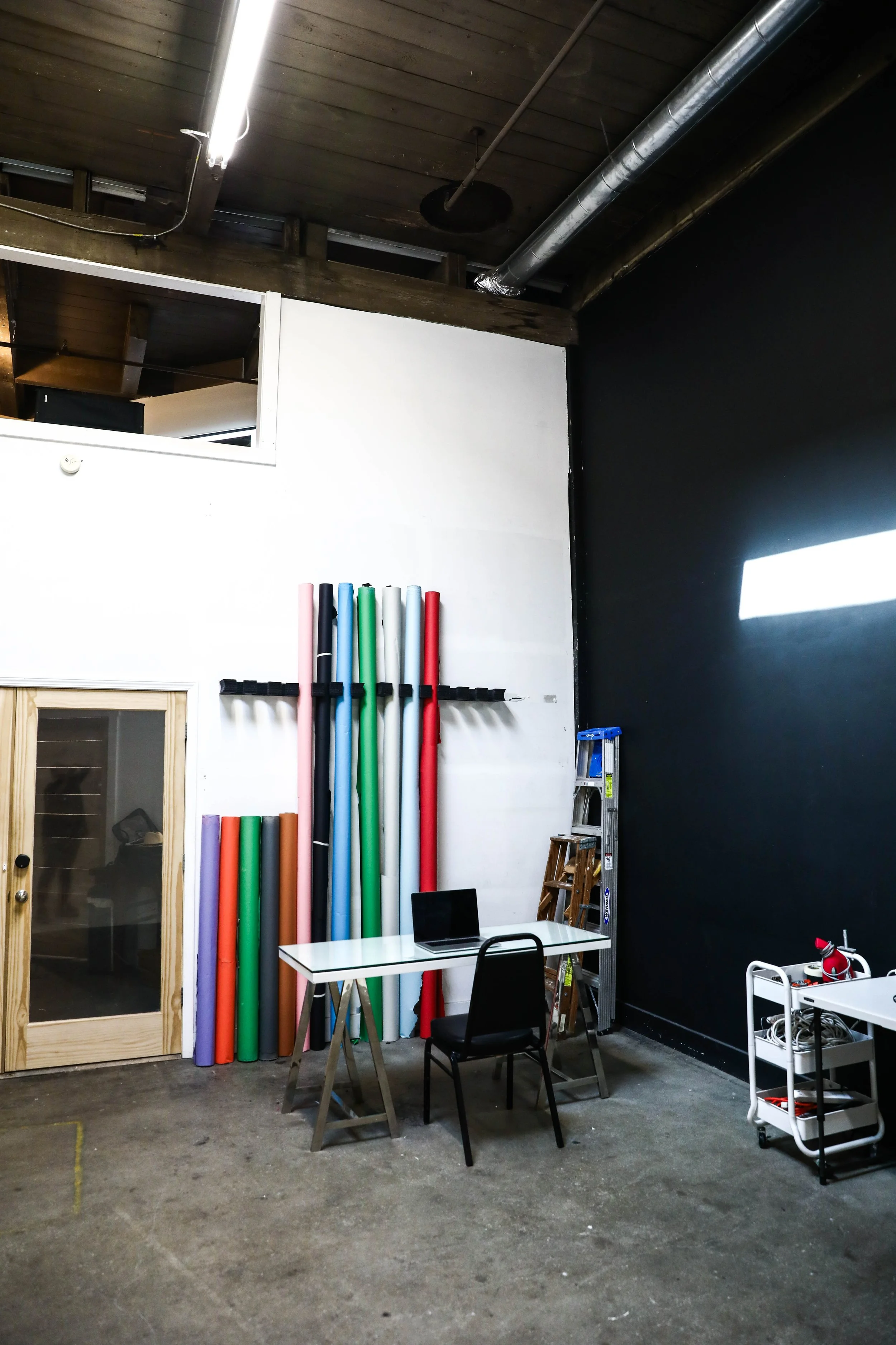 Industrial workspace with rolled colored paper, a white desk with a laptop, a black chair, a silver ladder, and a storage cart. The room has a concrete floor, black and white walls, and a wooden ceiling with visible pipes and lighting.