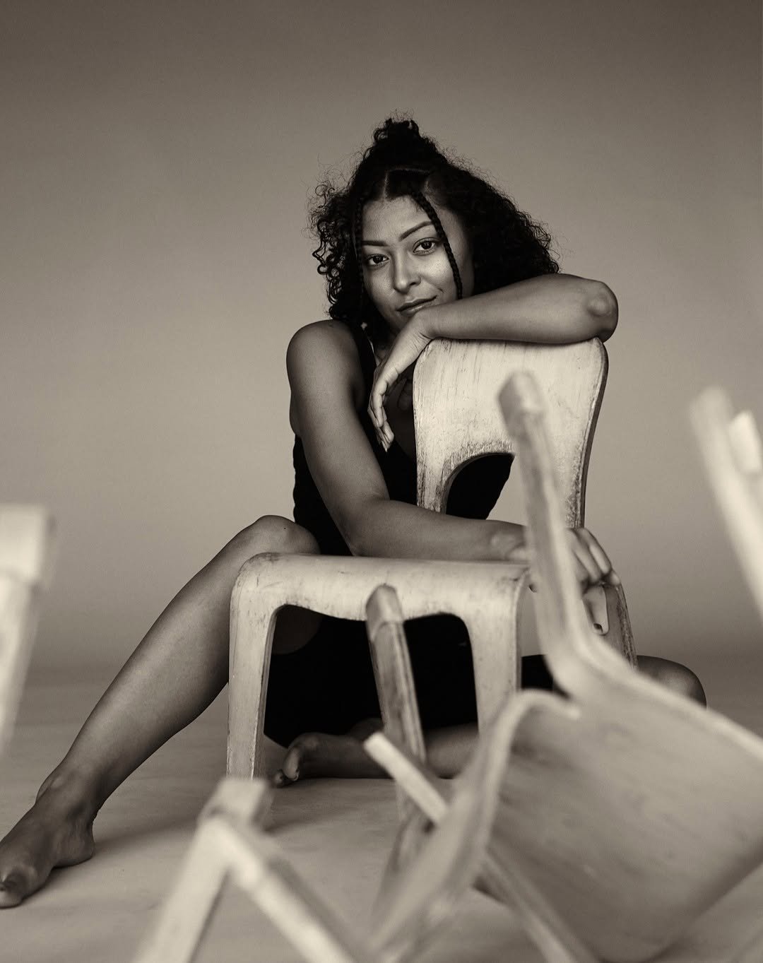 A black-and-white photo of a woman with curly hair, sitting behind a wooden chair, resting her head on her arm, with additional chairs scattered in the foreground.