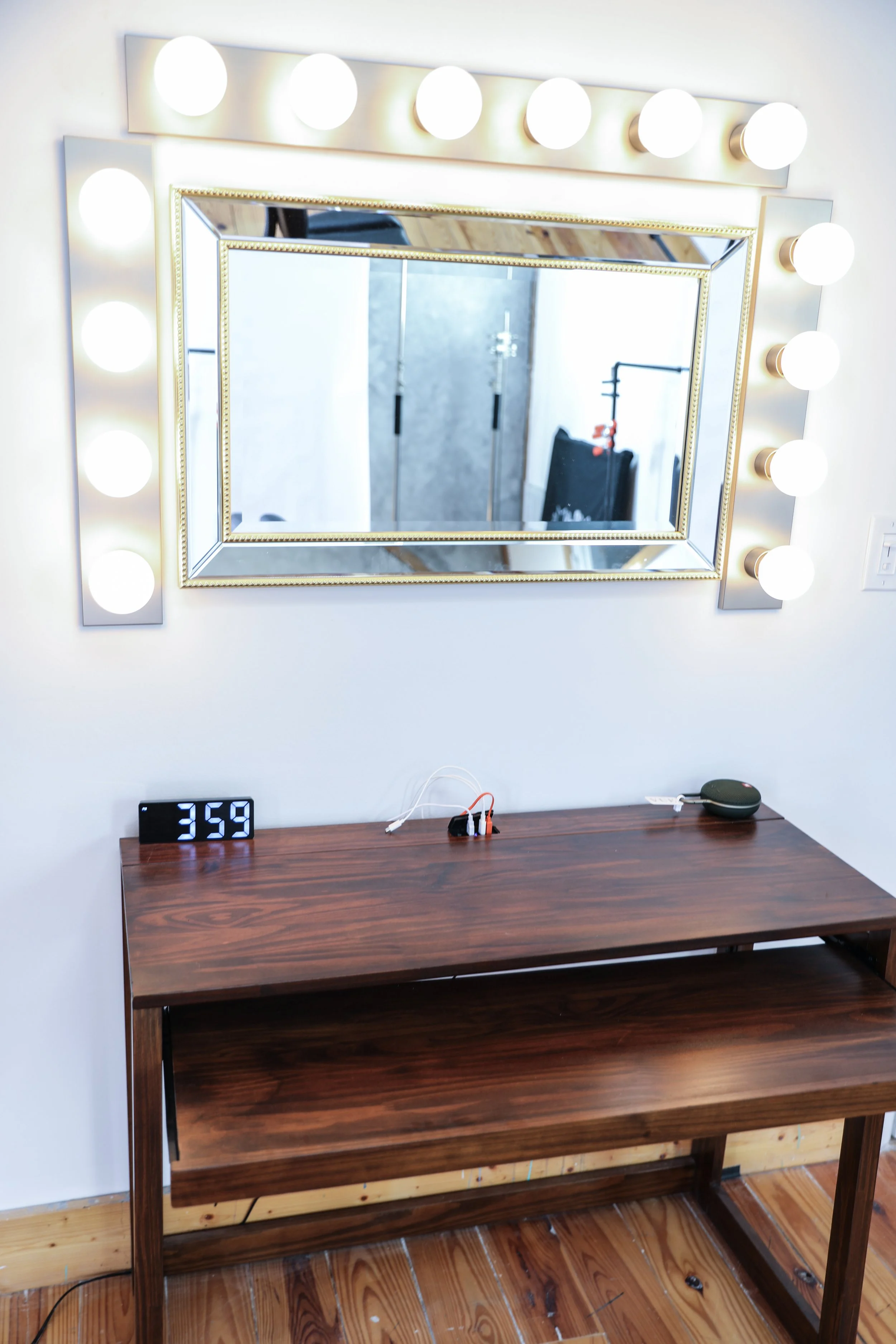 A large rectangular vanity mirror with Hollywood-style lights on each side hangs above a wooden table. On the table is a digital clock showing 3:59, some wires, and a small black device. The background shows a reflective surface of the mirror and par