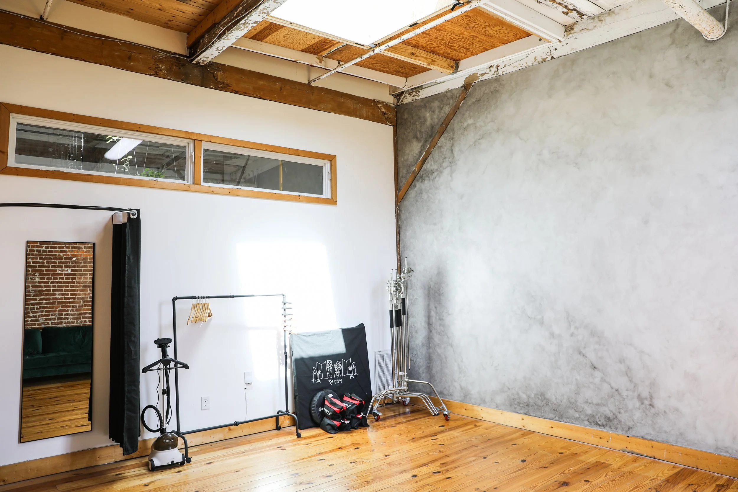 An empty room with wooden floors, a white wall with a mirror, suit bags, a folding clothes rack, and a large gray wall with patches of cement. There is a small horizontal window near the ceiling, and exposed wooden beams and ceiling panels.