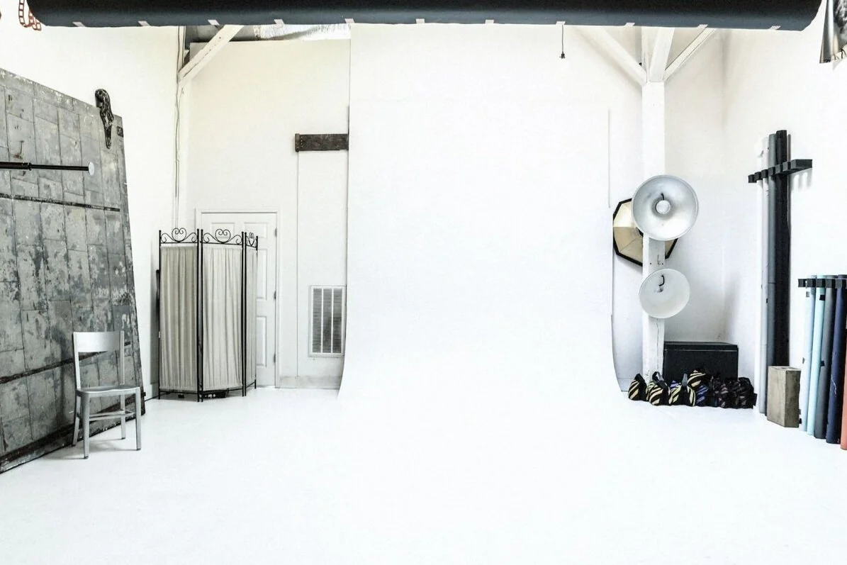 Photography studio with white backdrop, lighting equipment, and props including a chair, a folding screen, and rolls of colored paper.