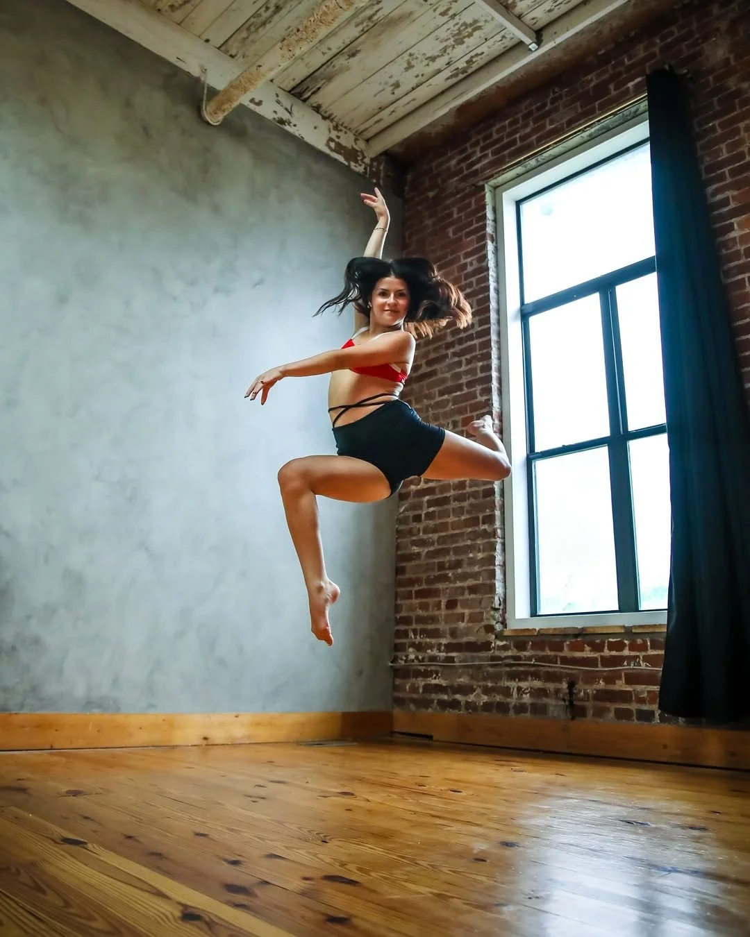 A woman jumping in a dance studio with wooden floors, brick and concrete walls, and a large window with black curtains.