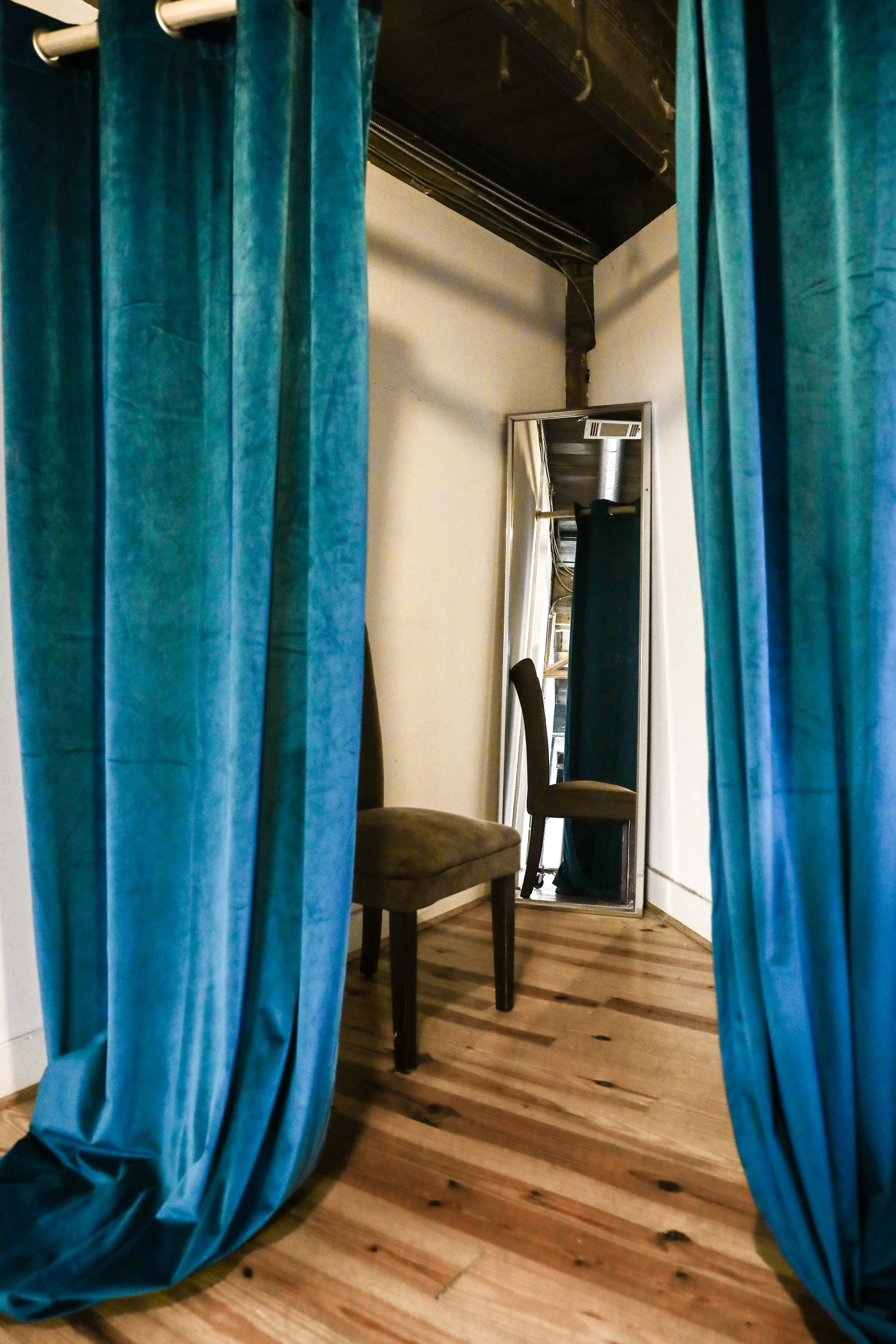 A fitting room with blue curtains, a wooden floor, a chair, and a tall mirror.