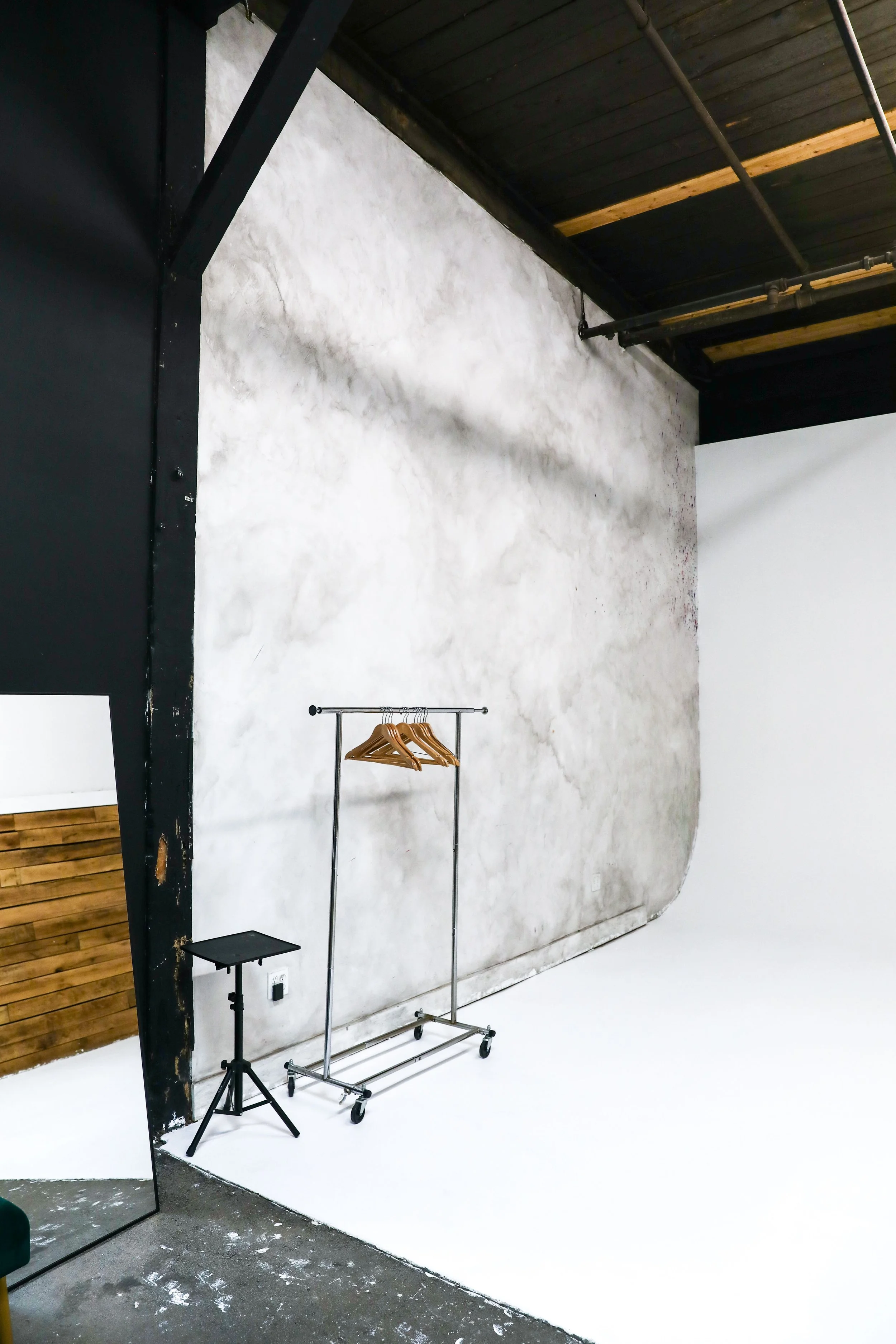 A photo of a photography studio with a weathered, textured gray backdrop, a metal clothing rack with wooden hangers, a small black tripod table, and a large mirror leaning against the wall.