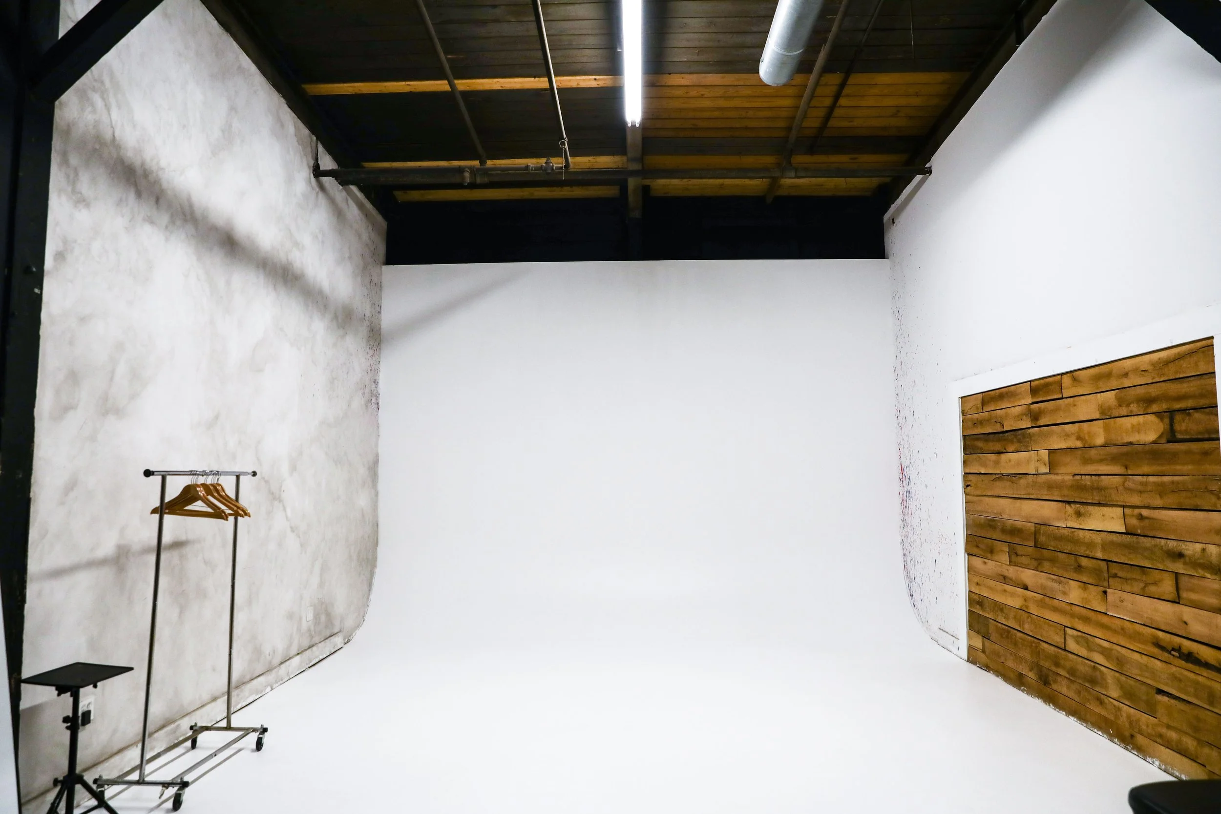 Empty photo studio with white backdrop, wood panel accent wall, clothing rack, and professional lighting equipment.