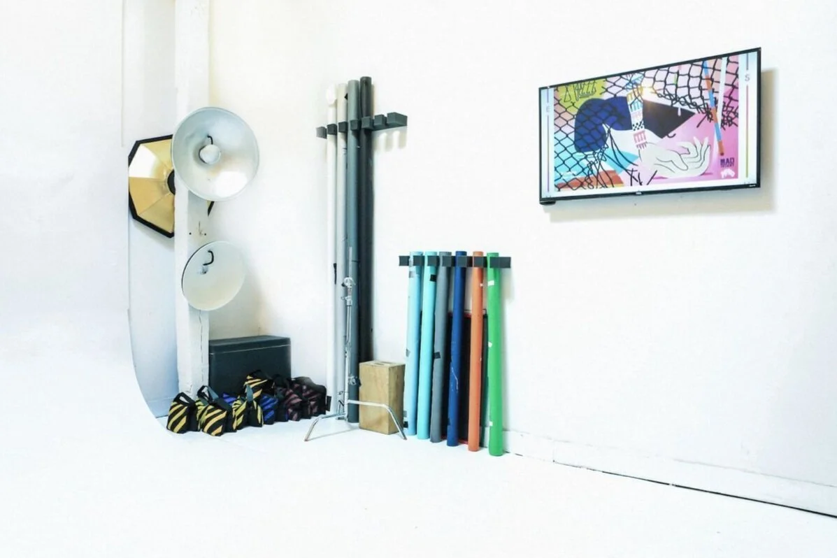 Interior of a photography or art studio showing lighting equipment, colored tubes, a painting on a wall, and photography accessories.
