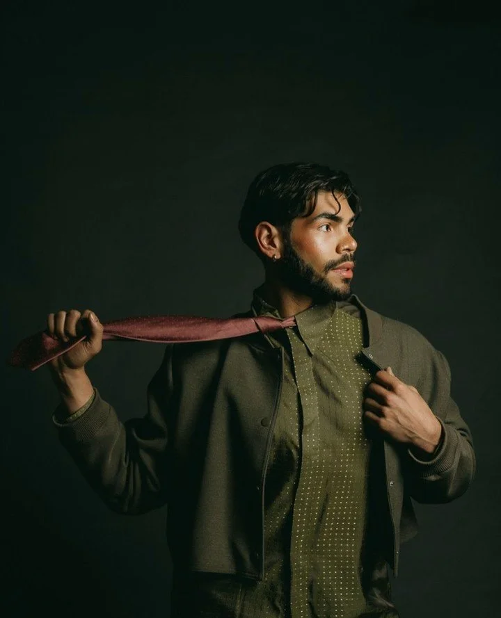 A young man with dark hair and beard holding a red tie over his shoulder, wearing a green patterned shirt and dark jacket, looking to the right against a dark background.