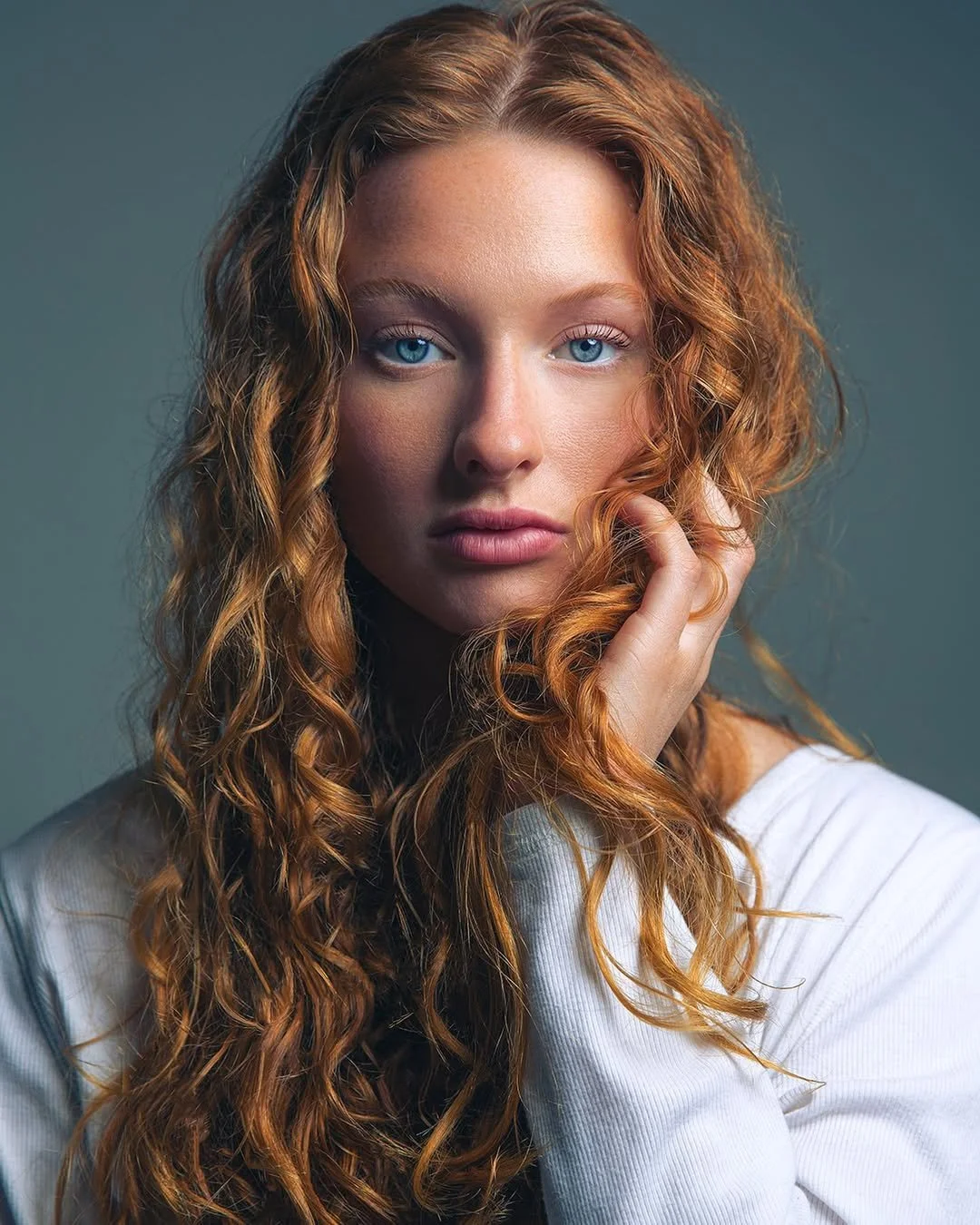 Close-up portrait of a woman with red curly hair and blue eyes, wearing a white shirt, touching her face with her right hand, against a plain muted background.