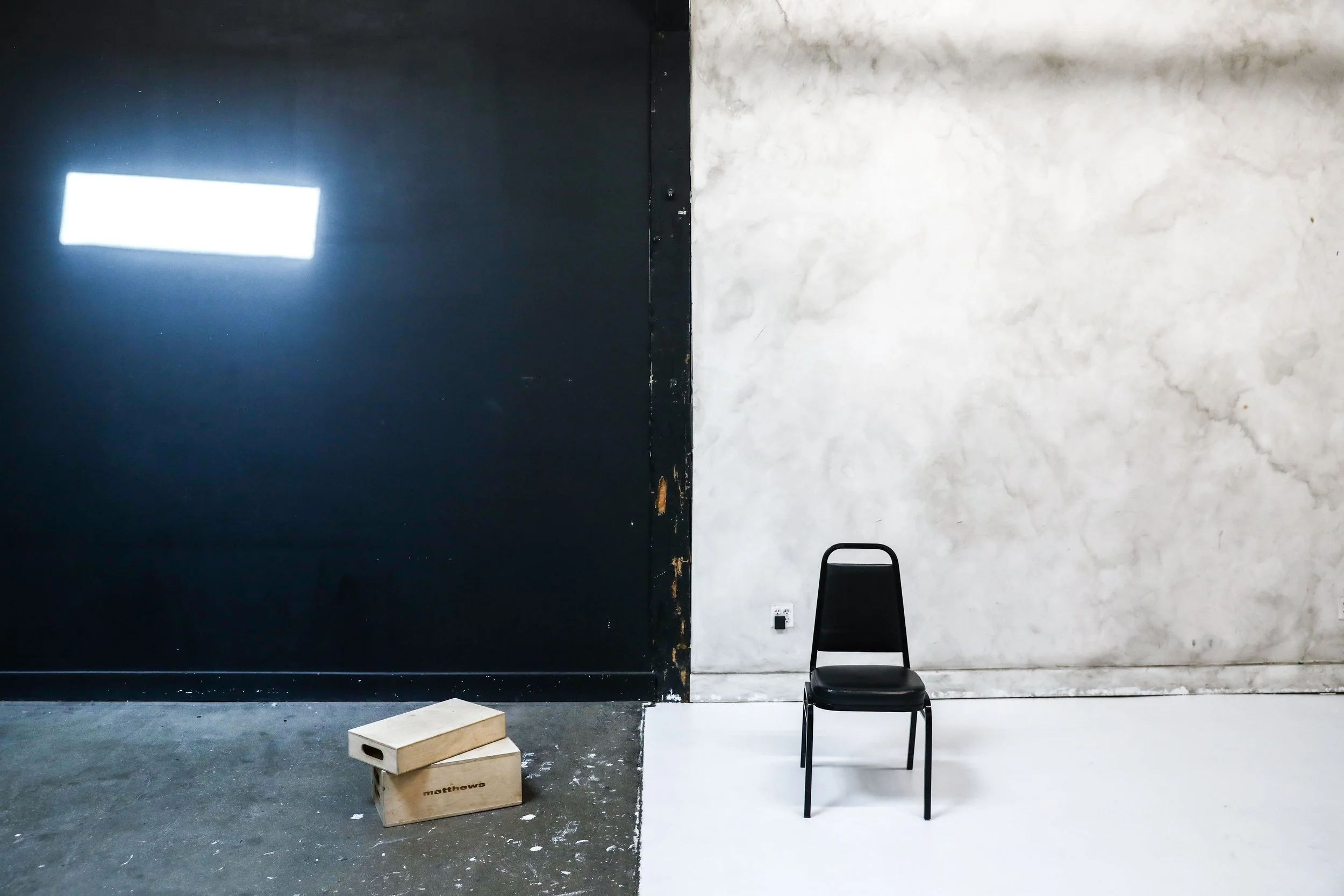 A black chair is positioned against a wall, divided into two sections: a black painted wall on the left and a white, textured wall on the right. Two wooden boxes are on the floor near the black wall, and there's a black electrical outlet on the white