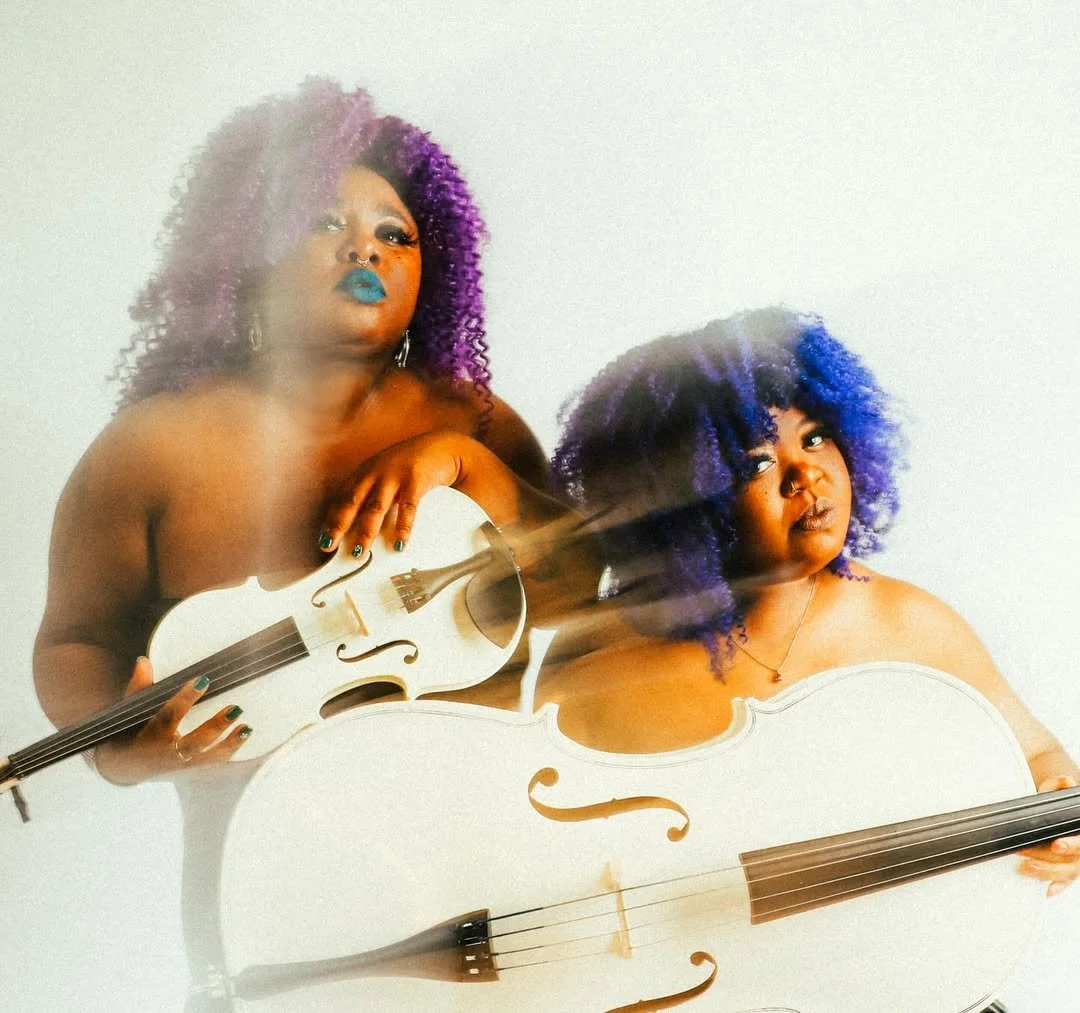Two women with purple and blue curly hair holding white violins, posing confidently against a plain white background.