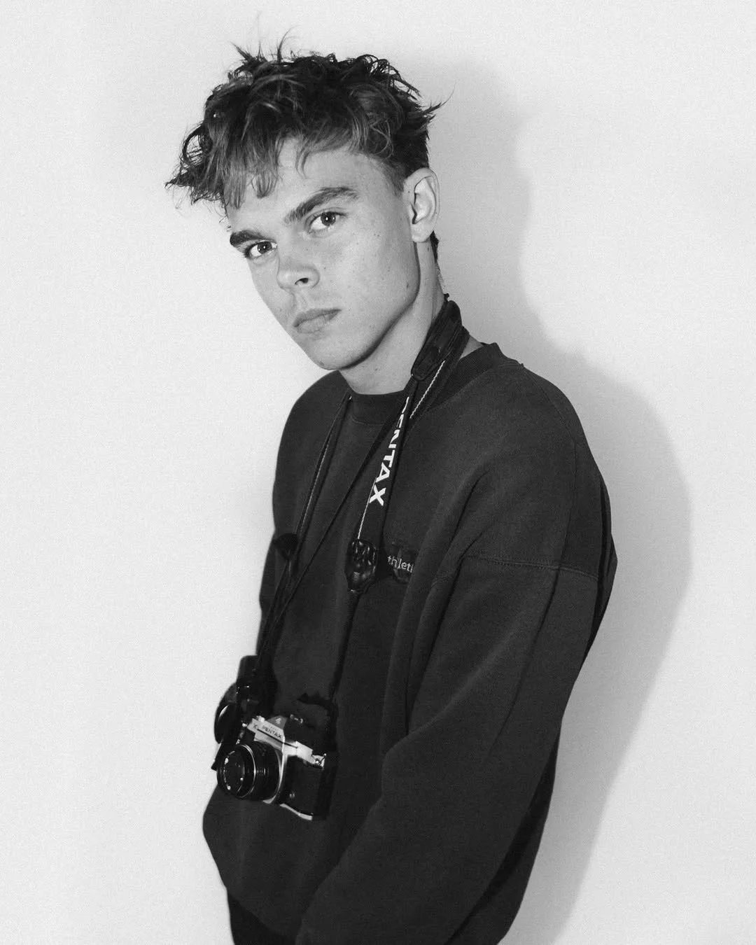 Black and white photo of a young man with messy hair, wearing a dark sweatshirt, with a camera hanging around his neck, standing against a plain white wall.