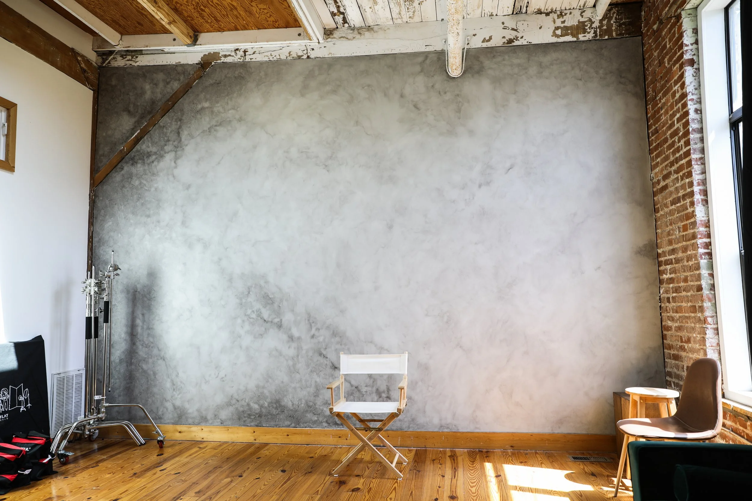 Empty room with wooden floor, a gray textured wall, brick wall, large window, and minimal furniture including a director's chair, a brown chair, and a small wooden table.