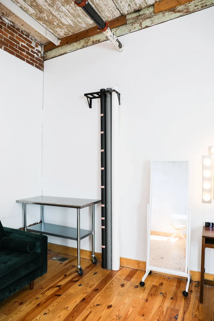 A corner of a room with a mirror on wheels, a small metal table, and three rolled-up posters leaning against a white wall.
