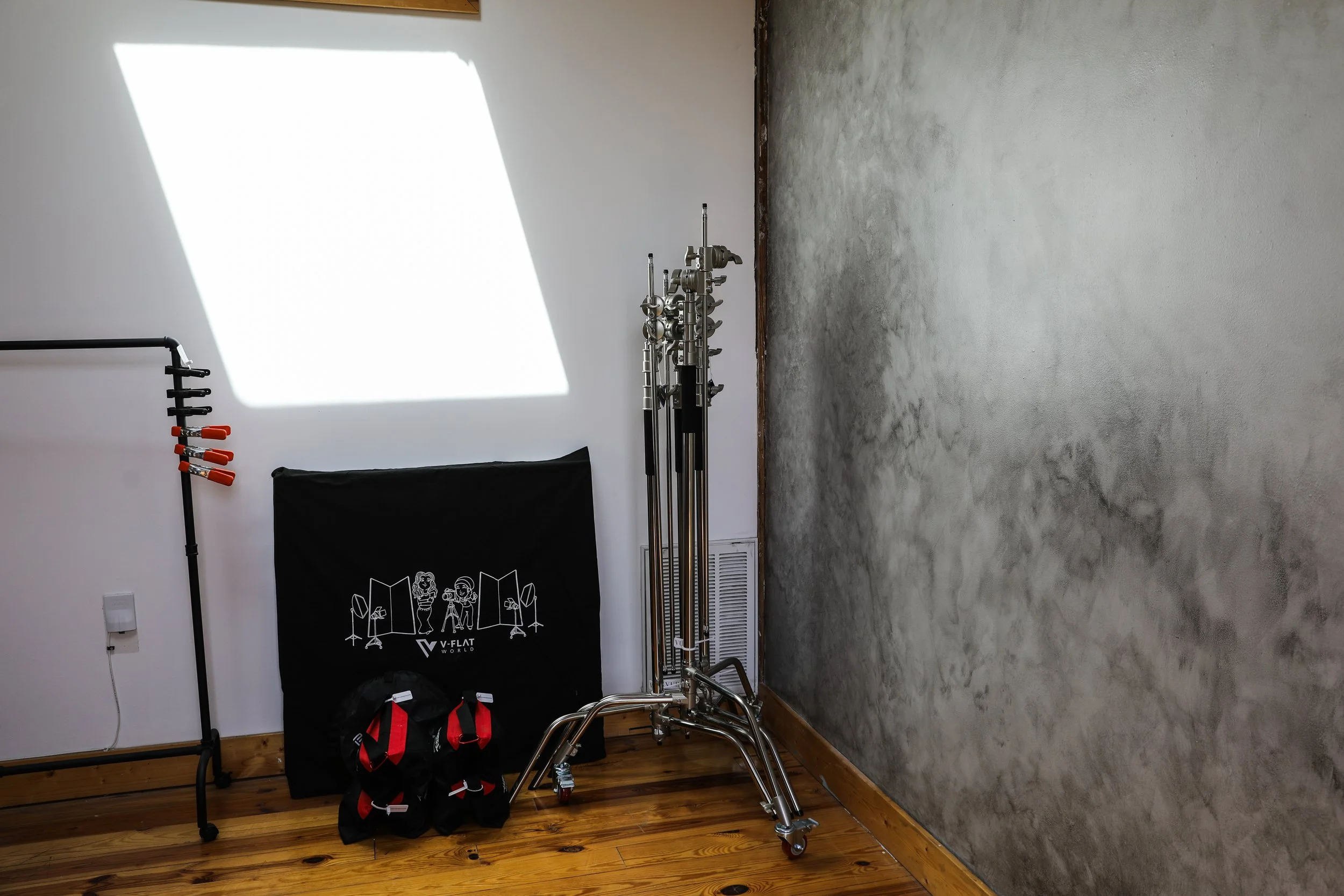 Photography studio setup with lighting equipment, a black backdrop, and weights, on a wooden floor and a wall with a textured plaster finish.
