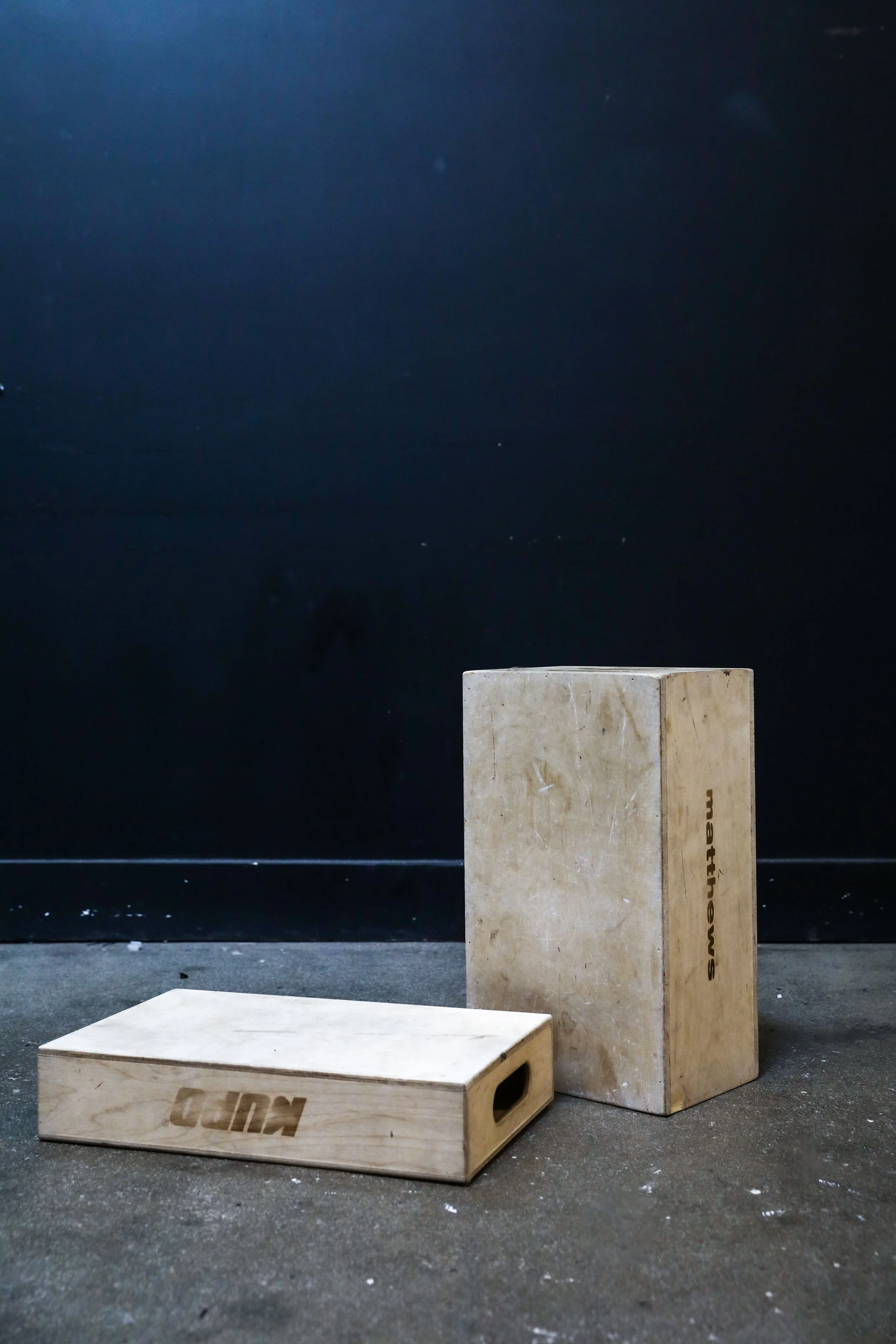 Two wooden plyometric jump boxes, one large and one small, placed on a concrete gym floor against a black wall.