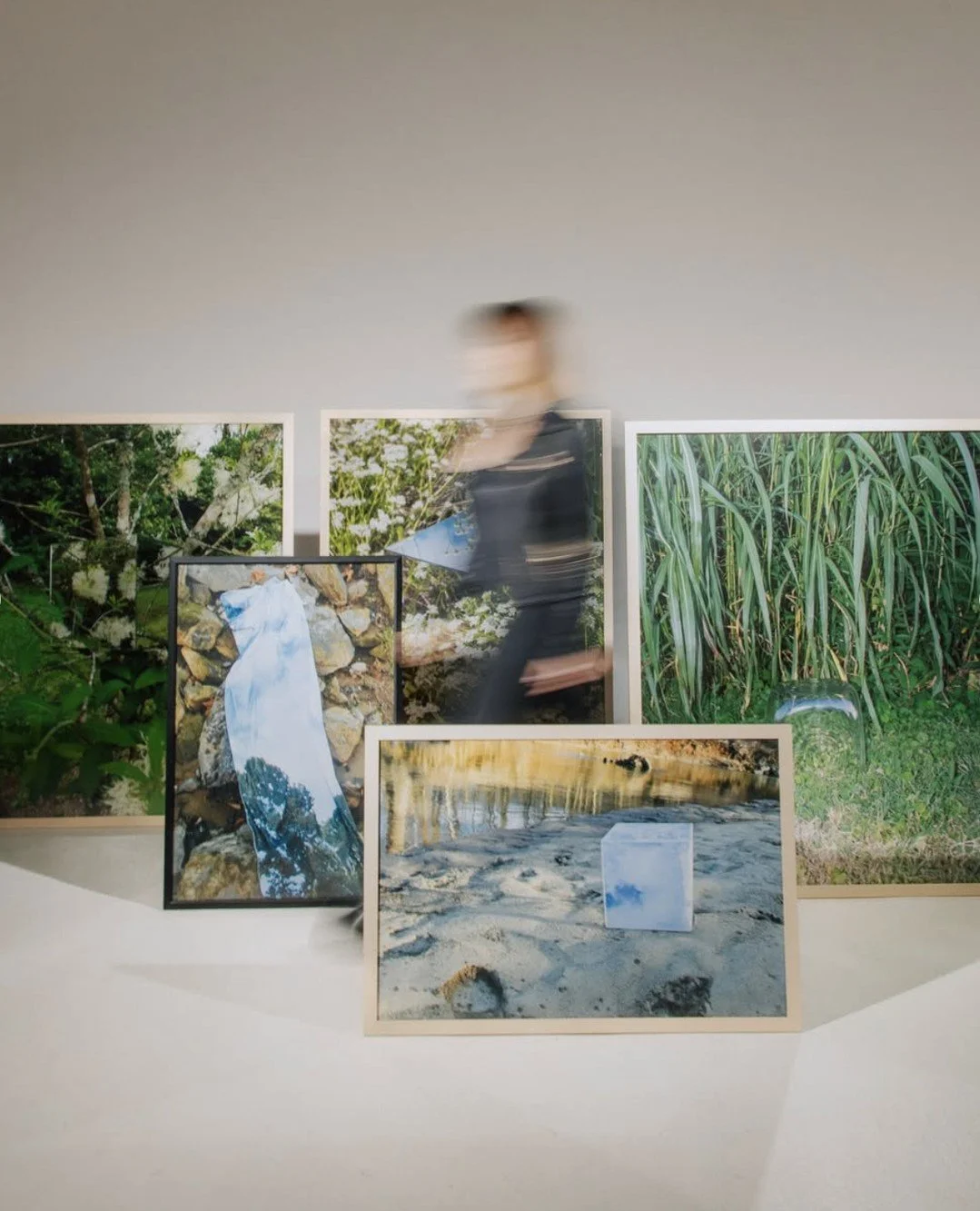 Blurry person walking past a display of nature photography images, including scenes of greenery, a river, and a white cloth on rocks.
