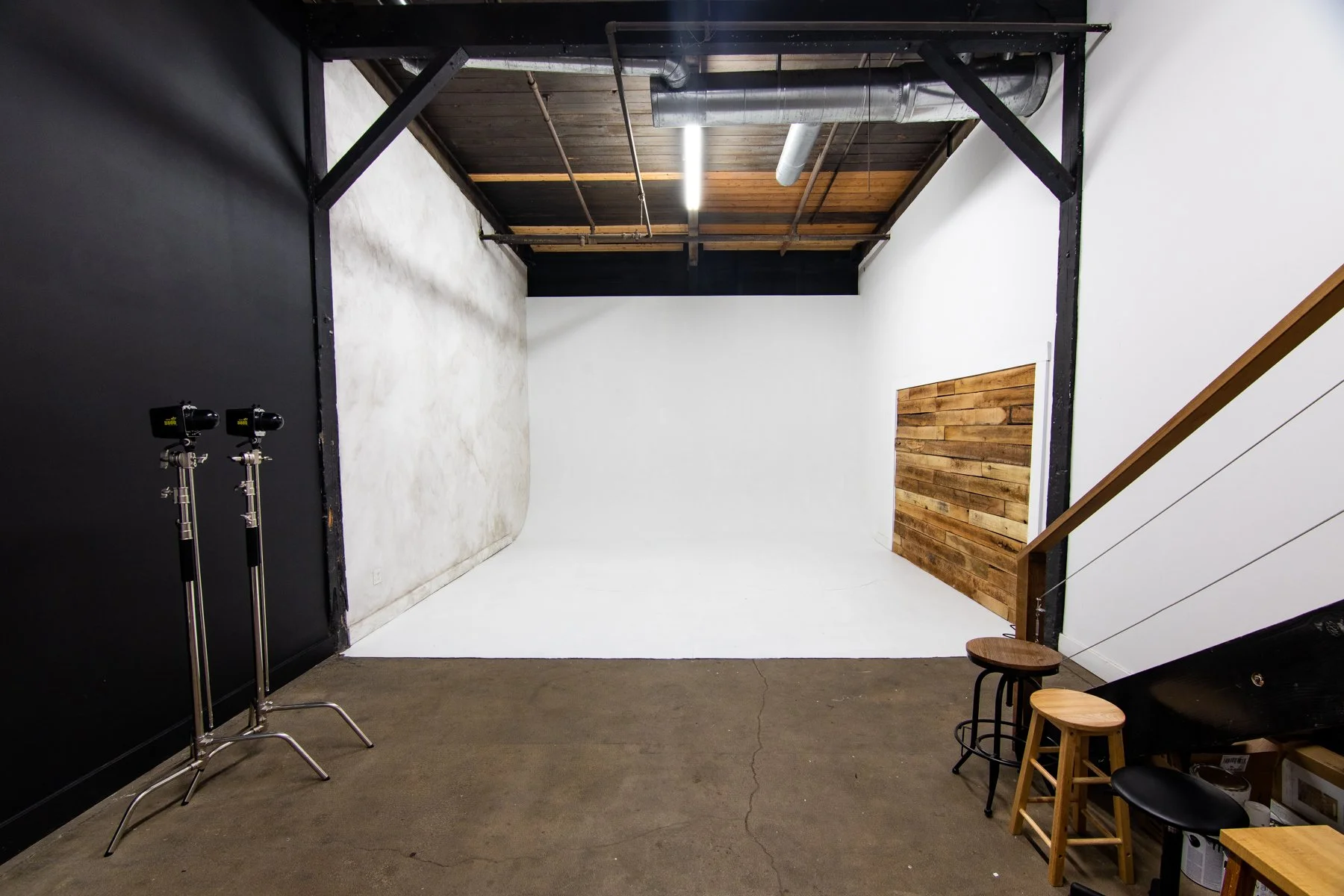 Photo of a photography studio with a white backdrop on the wall and a black wall on the left. There are two studio lights on stands on the left side, and the ceiling has exposed pipes and wooden beams. Stools and a small table are visible near the st