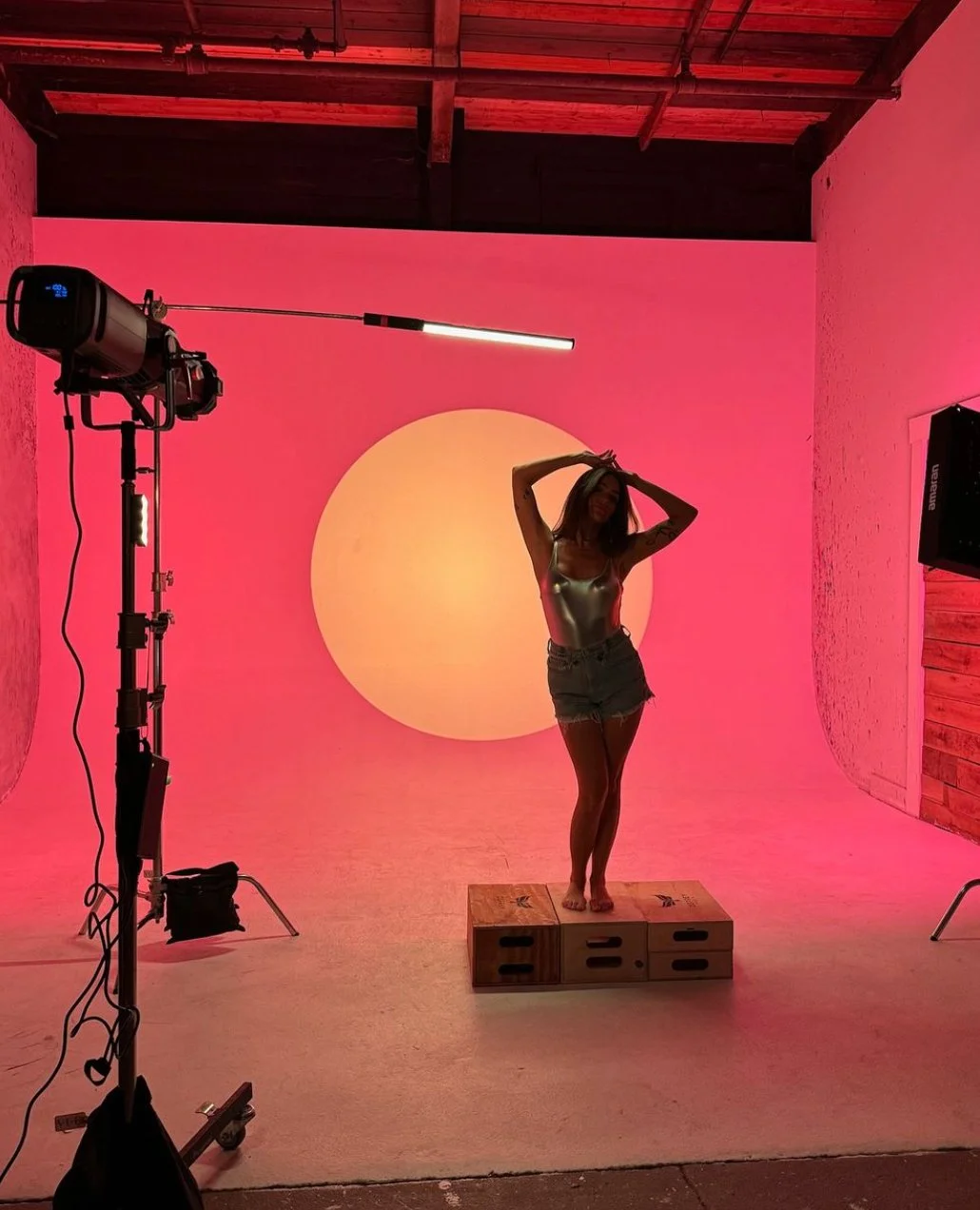 A woman posing on wooden crates in a studio with pink lighting, a large circular yellow sun projection on the wall behind her, and industrial lighting equipment nearby.