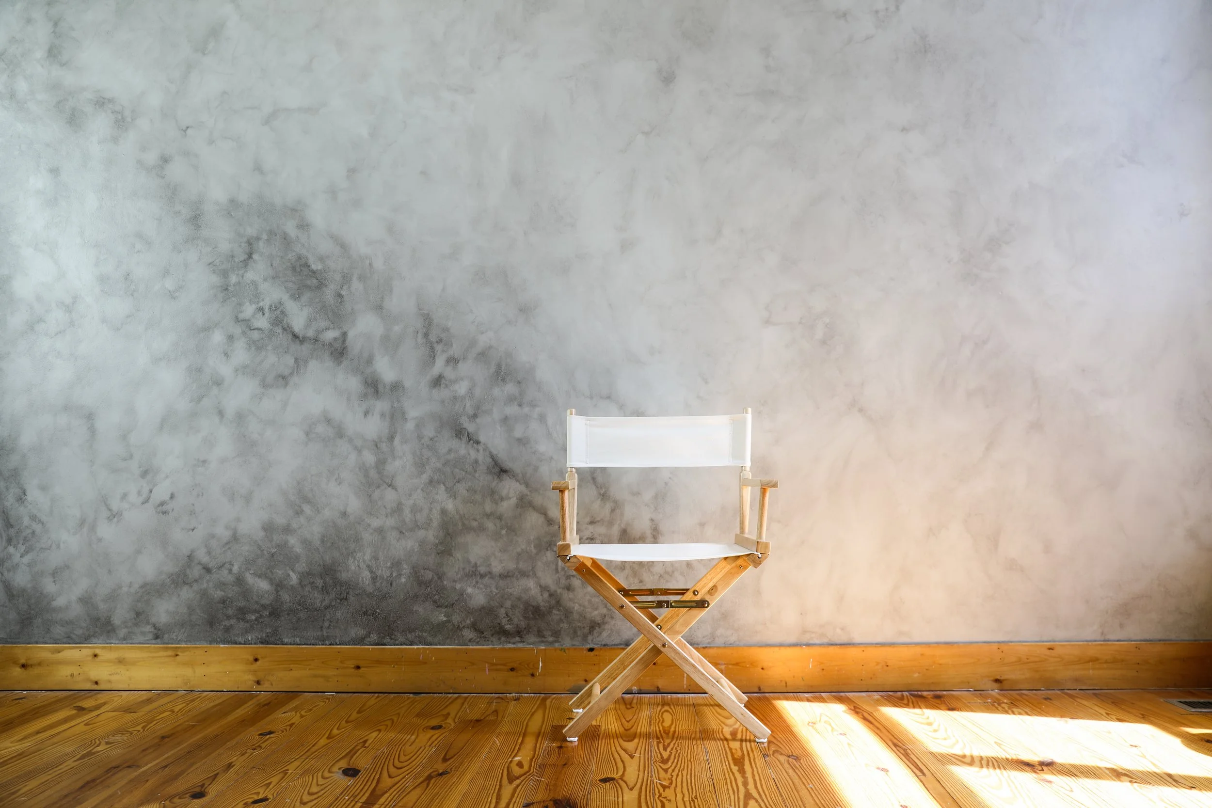 A director's chair with a white seat and backrest, and wooden armrests and legs, is positioned against a textured gray wall on a wooden floor with sunlight streaming in.